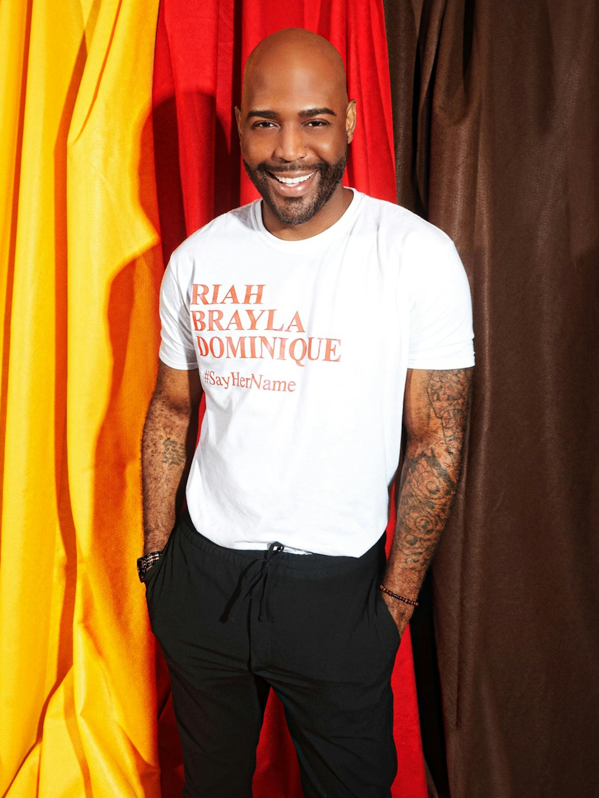 Karamo Brown wears a white t-shirt and black pants. His shirt reads: Riah Brayla Dominique #SayHerName. Behind him are yellow, red, and black hanging cloths.