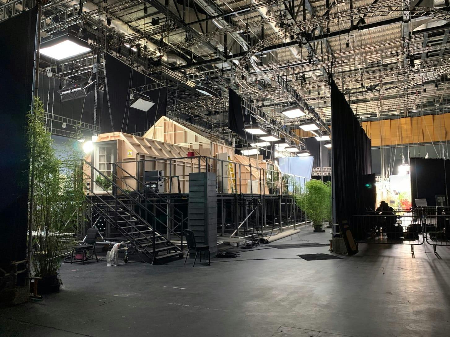 The soundstage used in Season 3 “Moments of Love” Master of None. There are stars, chairs, plants, curtains, overhead lights, and filming equipment surrounding the wooden house.