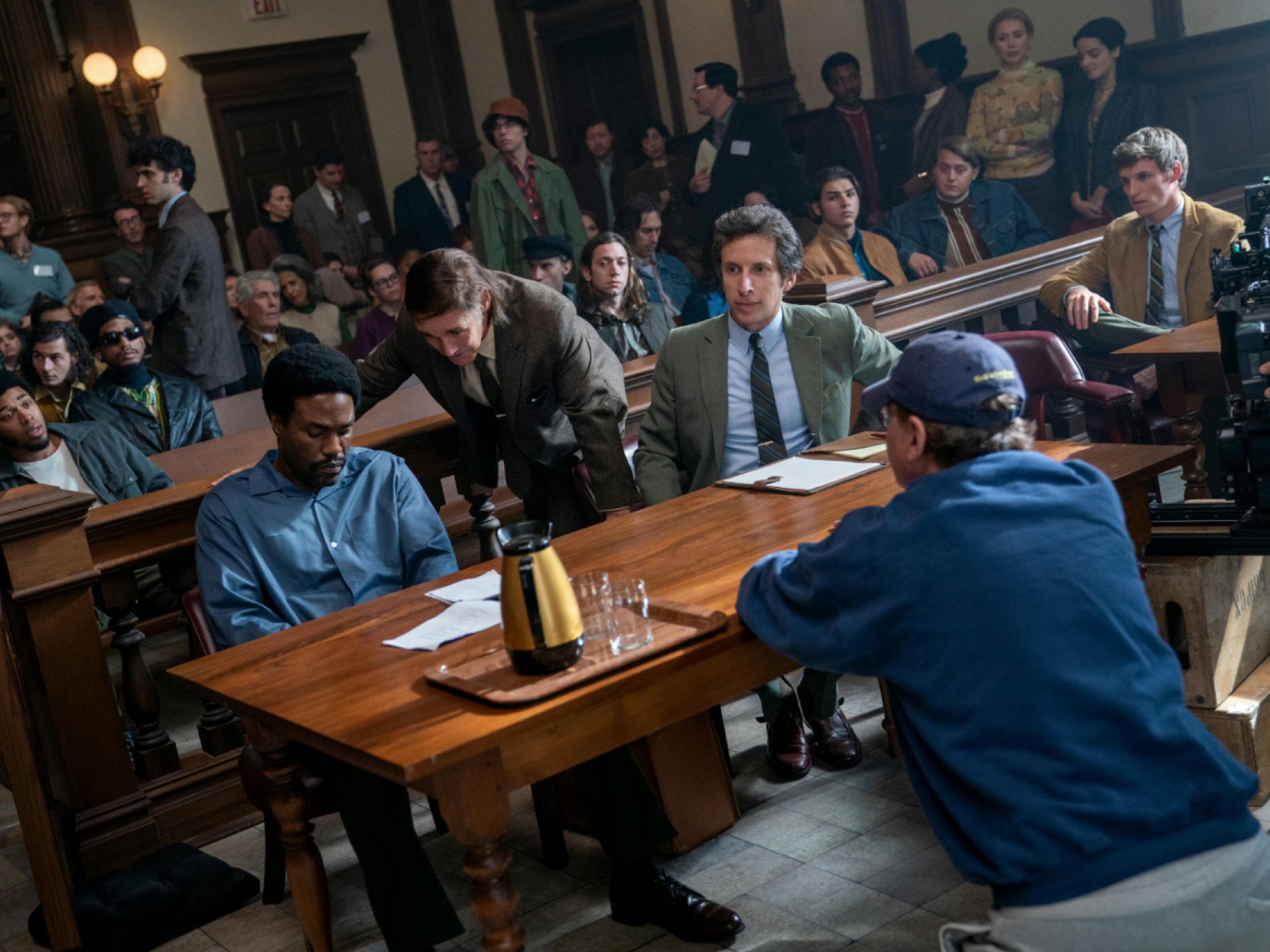 Abdul-Mateen, in costume as Bobby Seale, sits behind the defense table on this courtroom set, looking downwards. Fincher is pictured on the other side of the table, kneeling down. Mark Rylance, who plays lawyer William Kunstler, leans over Abdul-Mateen’s shoulder. Behind them extras fill the courtroom, as well as several actors portraying members of the Black Panthers.