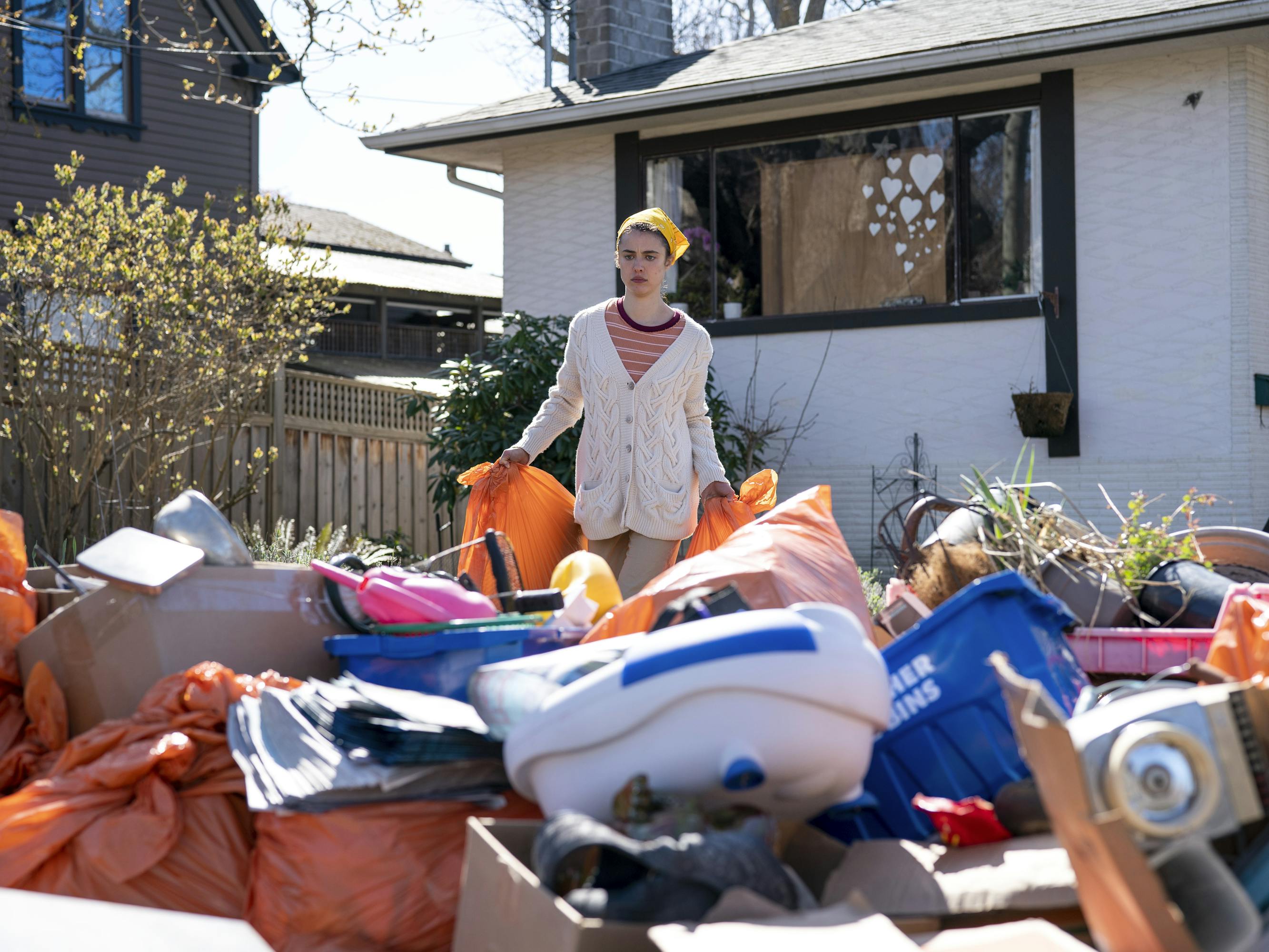 Alex Russell (Margaret Qualley) stands amongst a yard full of junk. She wears an oatmeal cardigan and a yellow bandana.