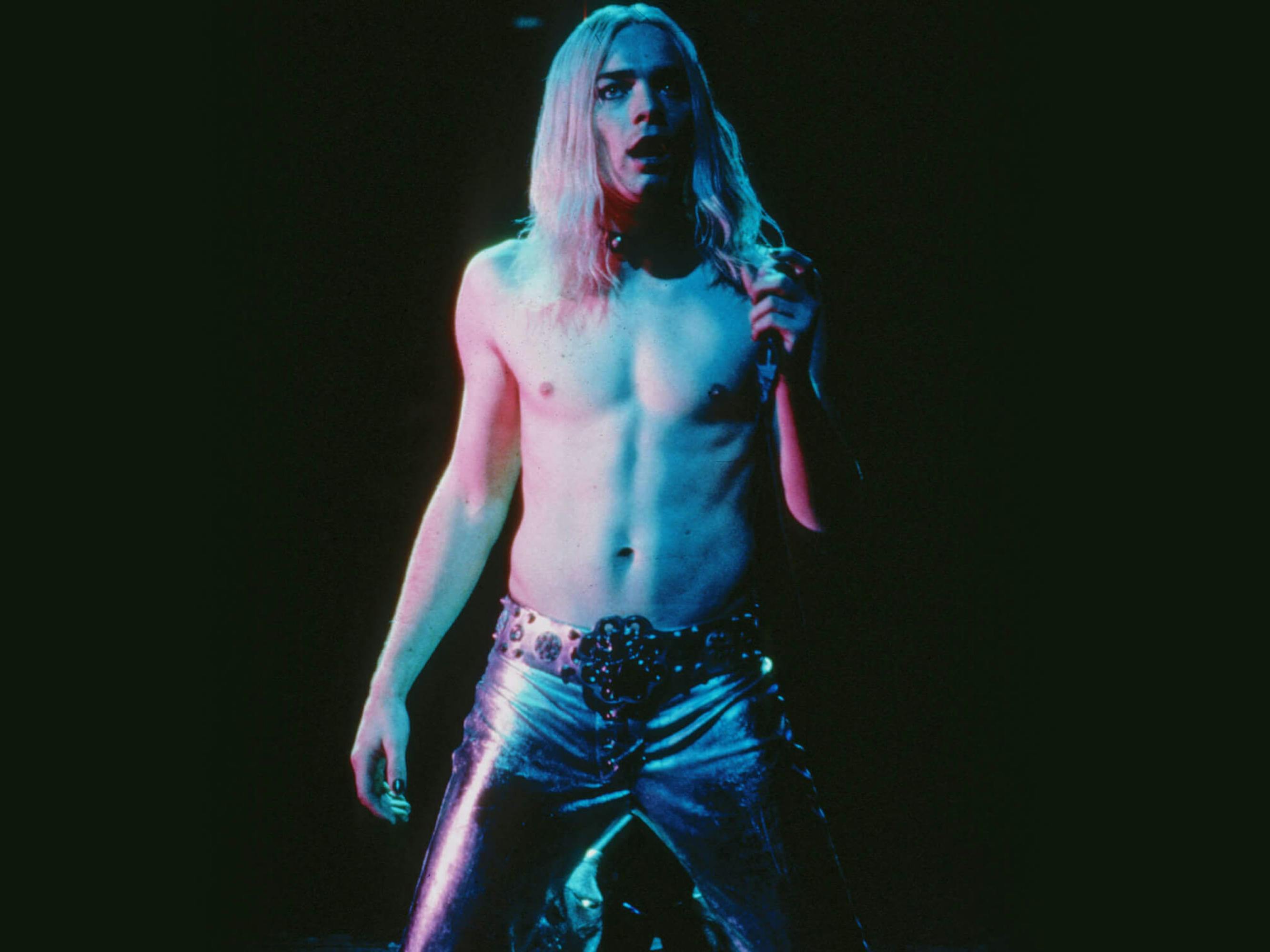 Curt Wild (Ewan McGregor) in Velvet Goldmine. The background is dark, accentuating his pale bare chest, long blond hair, and shiny pants.