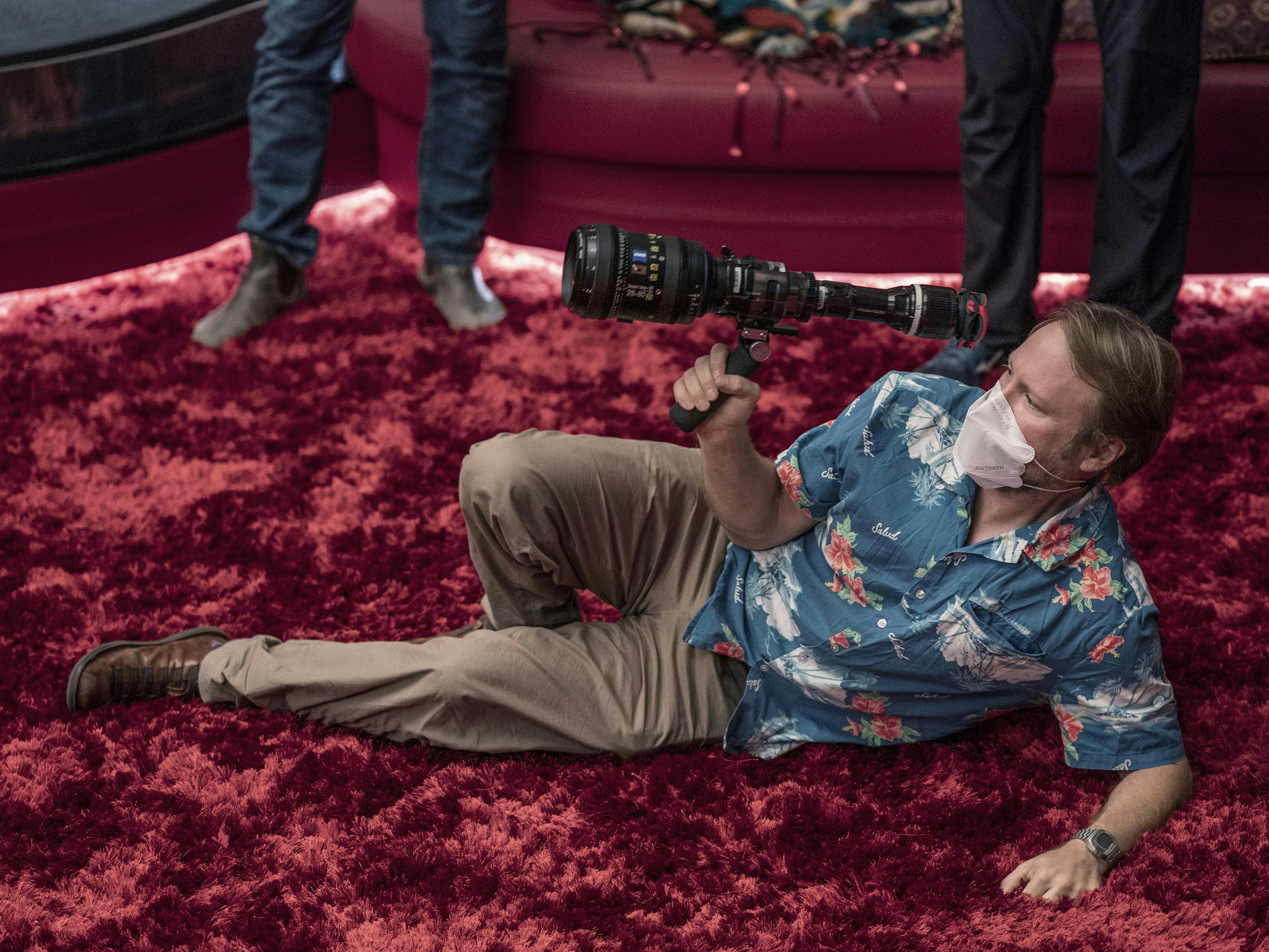 Rian Johnson wears a blue Hawaiian shirt and khakis and stretches out on a red velvet rug holding a camera.
