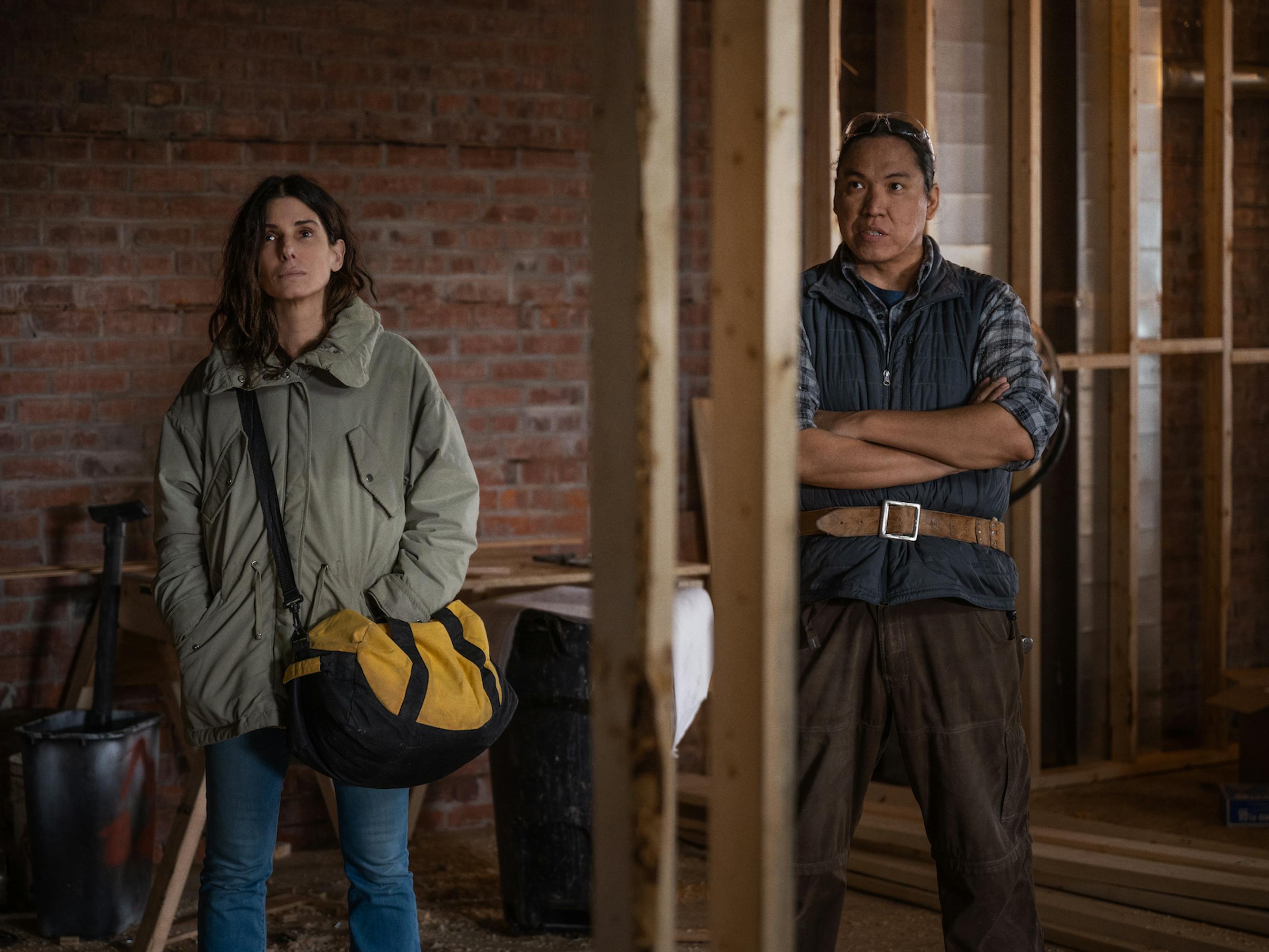 Ruth Slater (Sandra Bullock) and a fellow carpenter assess an empty room. Bullock wears a light green jacket and blue jeans, carrying a yellow duffel. The carpenter wears a checkered shirt, navy vest, and brown pants. 