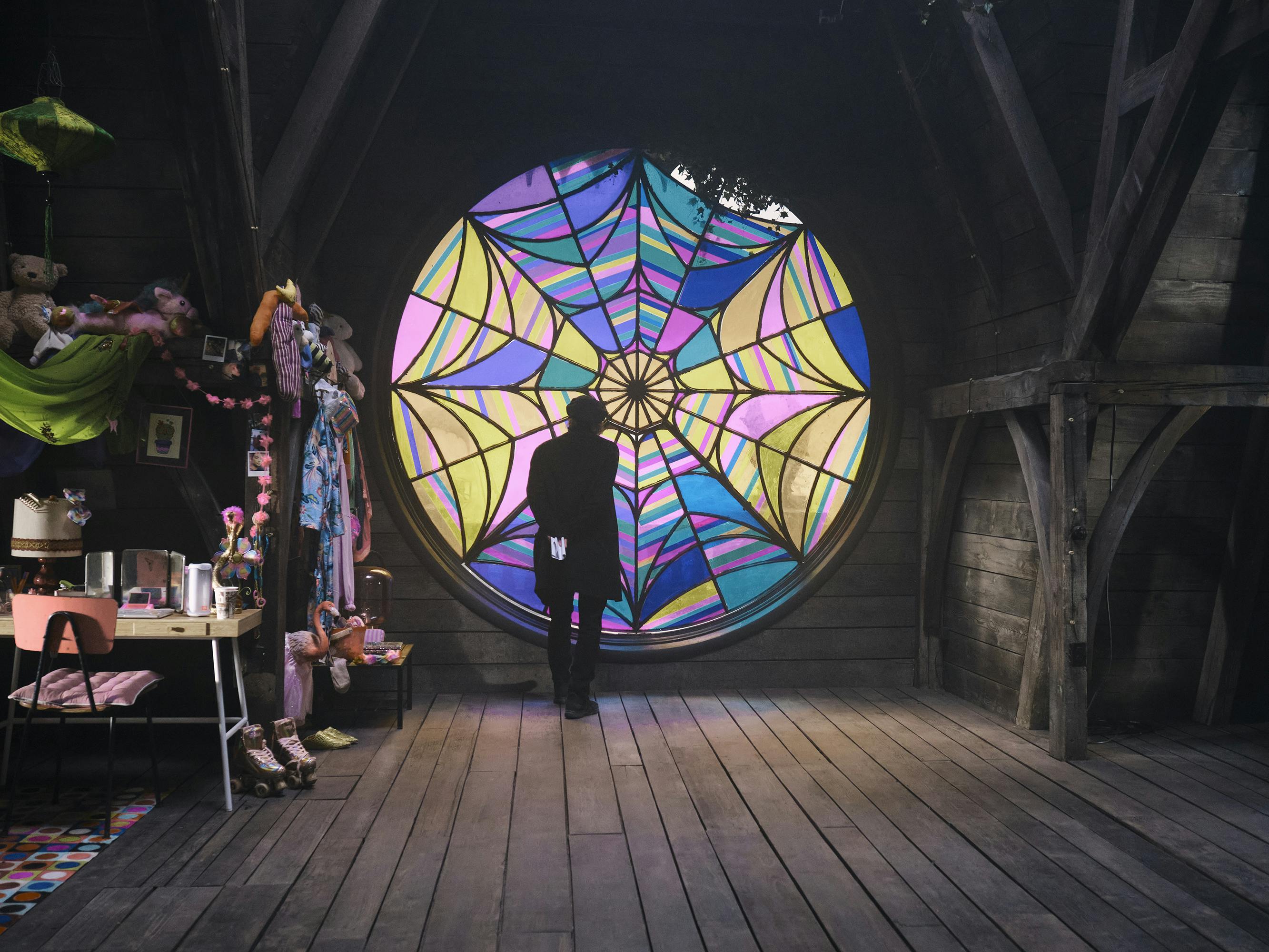 Tim Burton stands with his back to the camera, looking at the colorful stained glass window in Enid and Wednesday’s room.