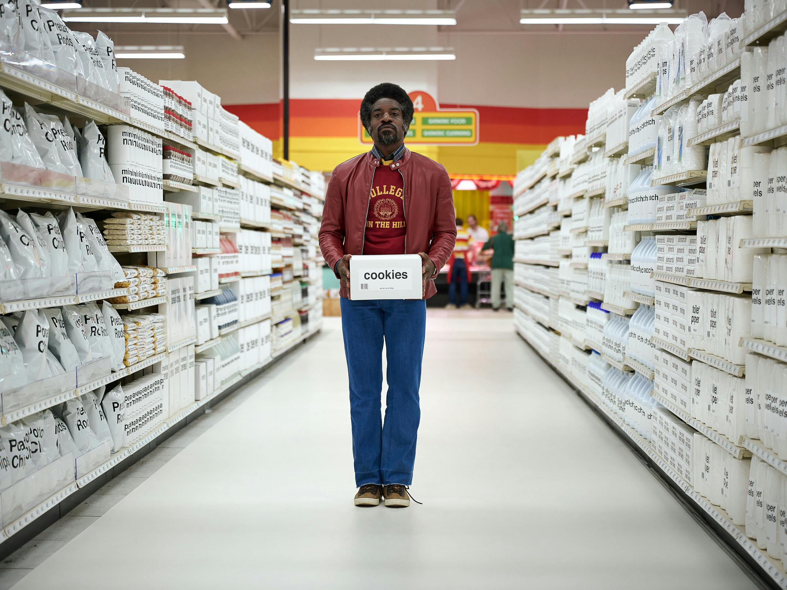 André Benjamin holds a white box of cookies and stands in an aisle of all-white products with black writing.
