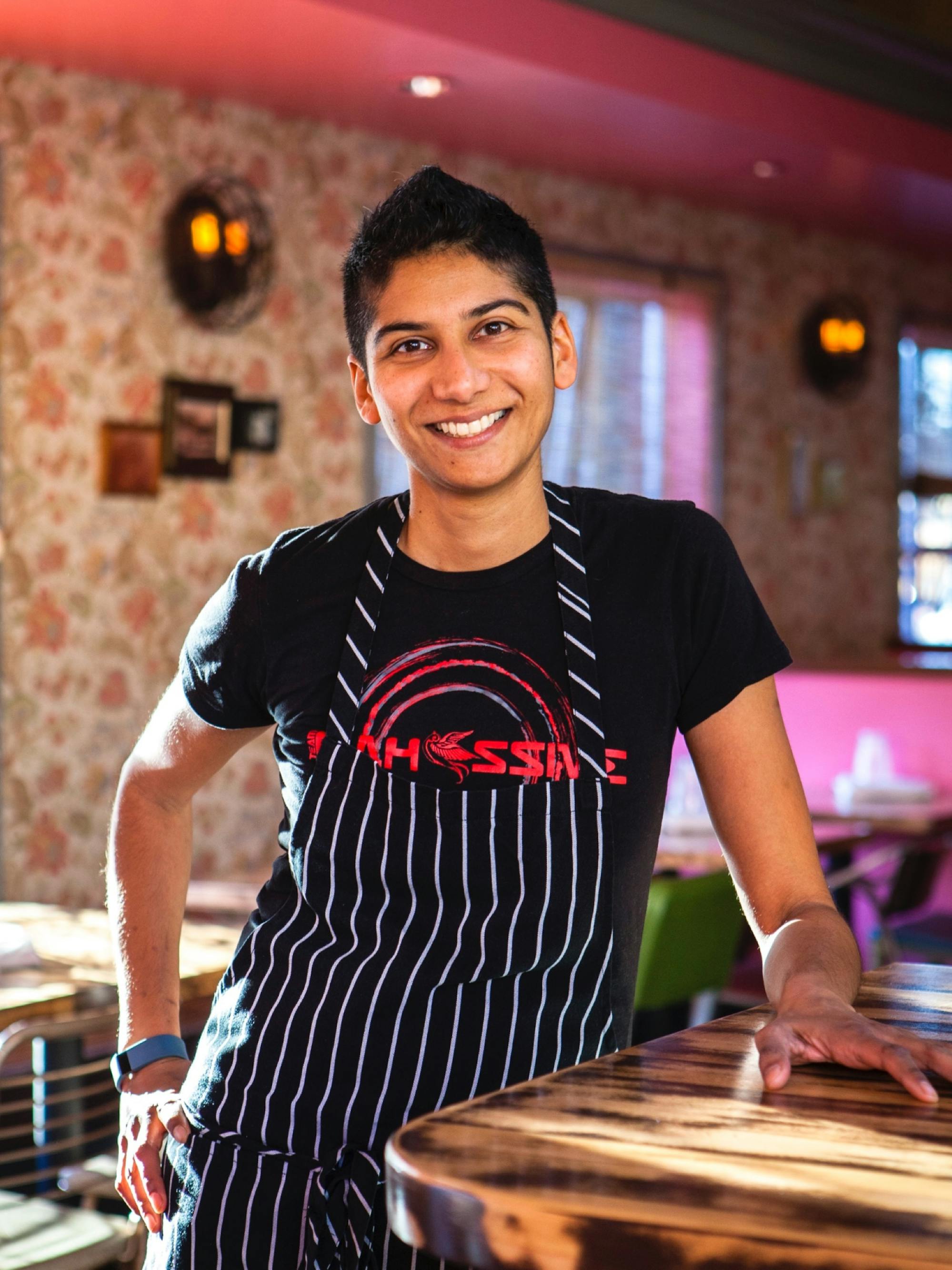Mistry at their Oakland restaurant, Juhu Beach Club. They’re smiling and leaning on a wood counter. They’re wearing a striped apron and black T-shirt with a red graphic. The restaurant features bright natural light, an exposed brick wall, and cheerful pink paint.