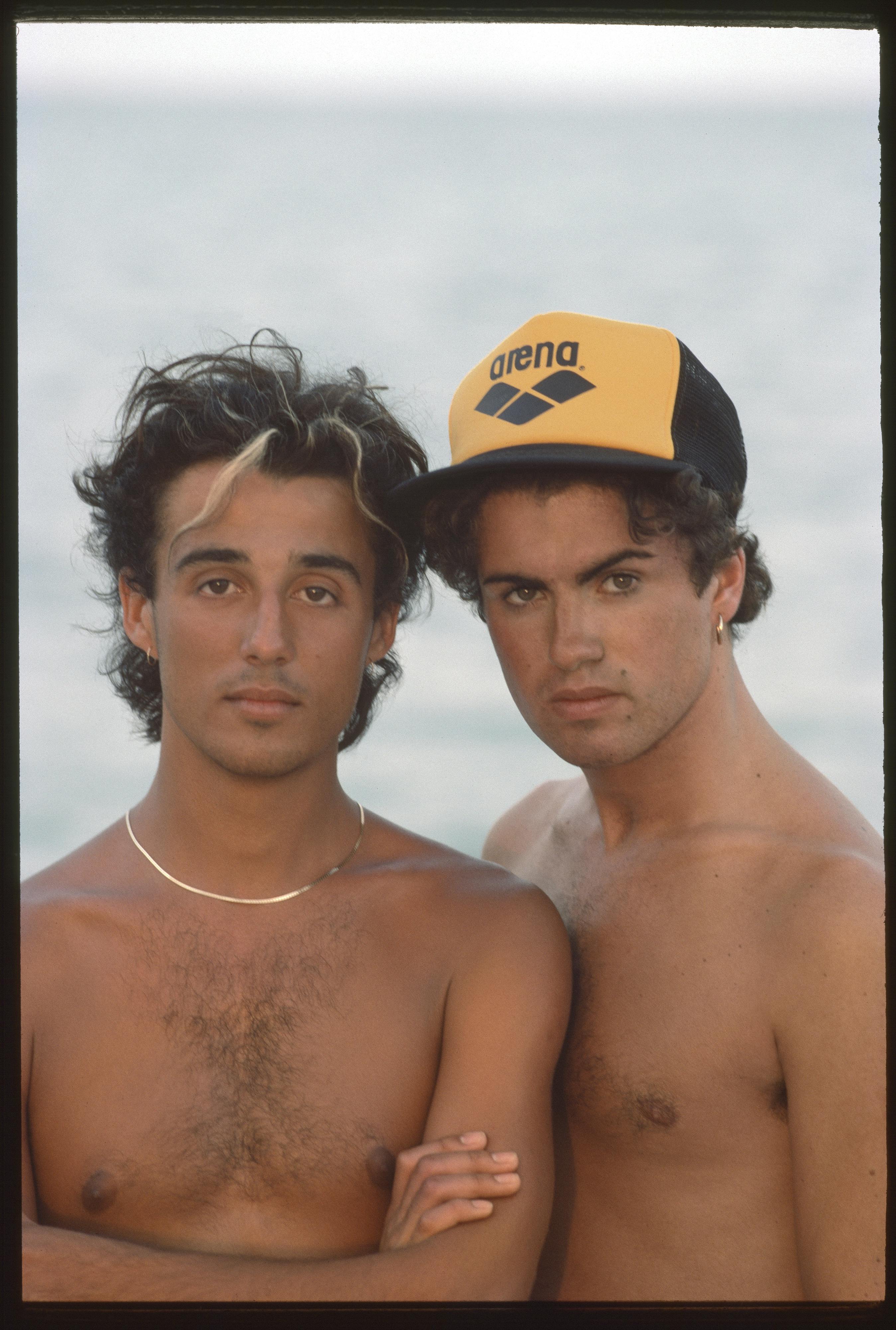 Andrew Ridgeley and George Michael pose shirtless against a beachy background.