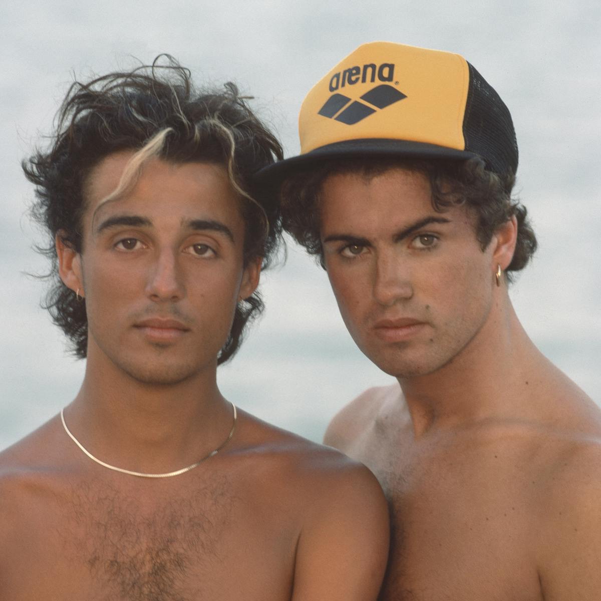 Andrew Ridgeley and George Michael pose shirtless against a beachy background.