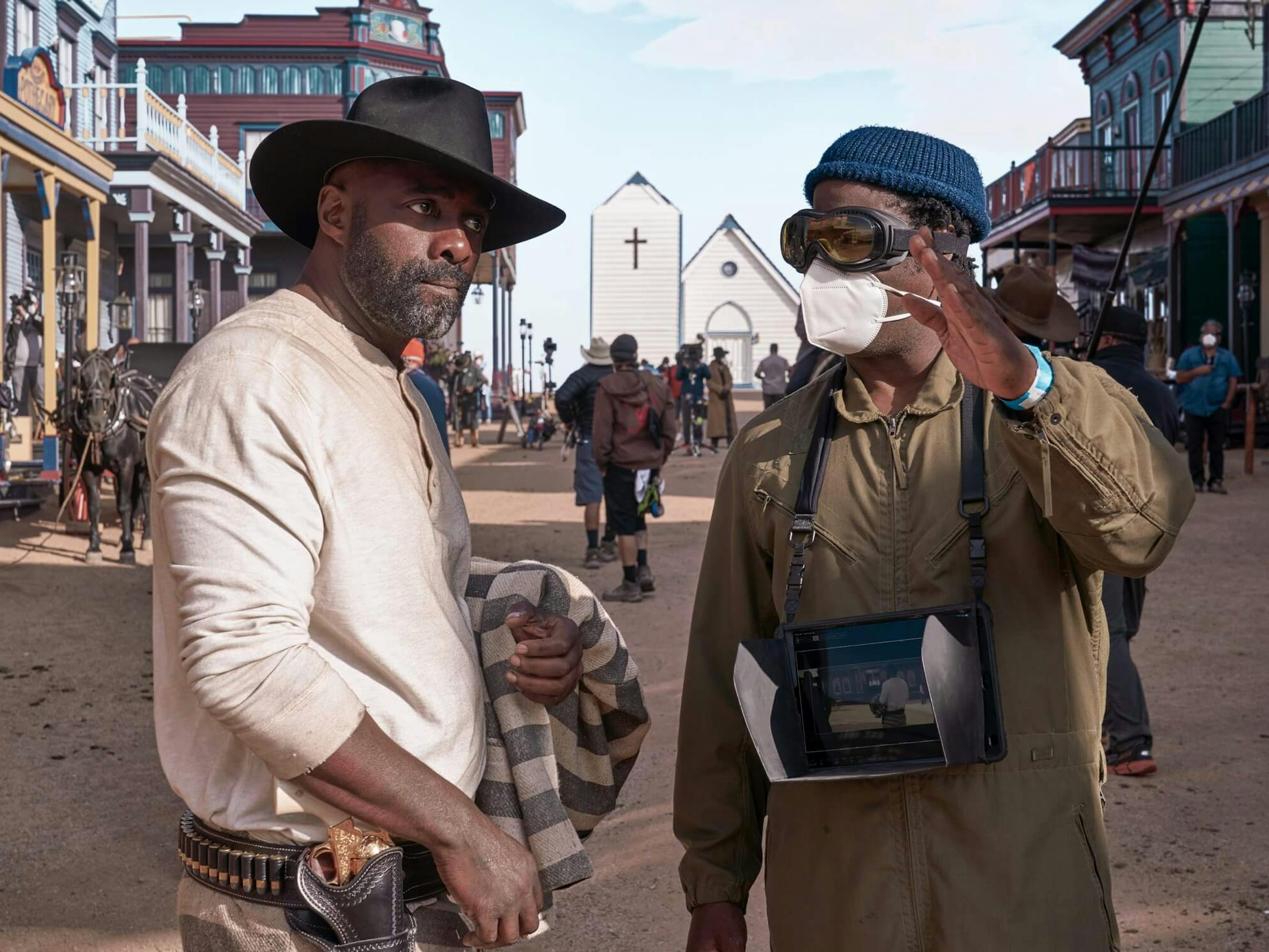 Idris Elba and Jeymes Samuel stand together on set. In the background is the chapel, and some storefronts. Elba wears a white shirt, dark hat, and carries his prison uniform in one arm. Samuel wears a tan jumpsuit and wears a teal beanie.