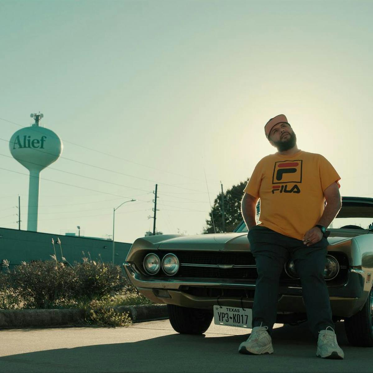 Mo Amer wears a yellow fila shirt and stands against his car. Behind him is a Alief water tower.