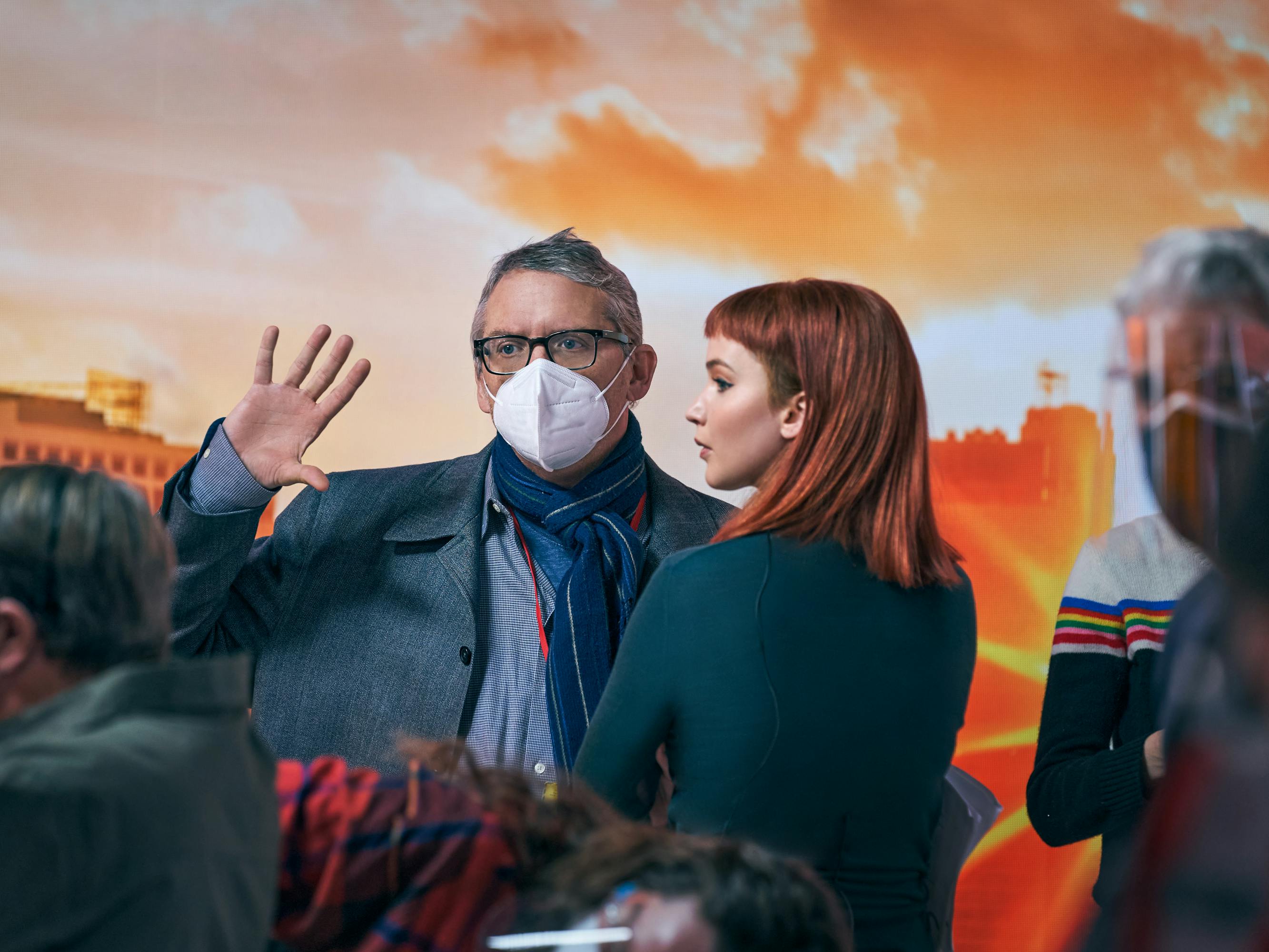 Adam McKay and Jennifer Lawrence stand on set. McKay wears a demin jacket, blue scarf, black glasses, and white mask. Lawrence wears a green shirt and has bright red hair. Other crew members mingle about in protective masks. Behind them is an orange-lit background.