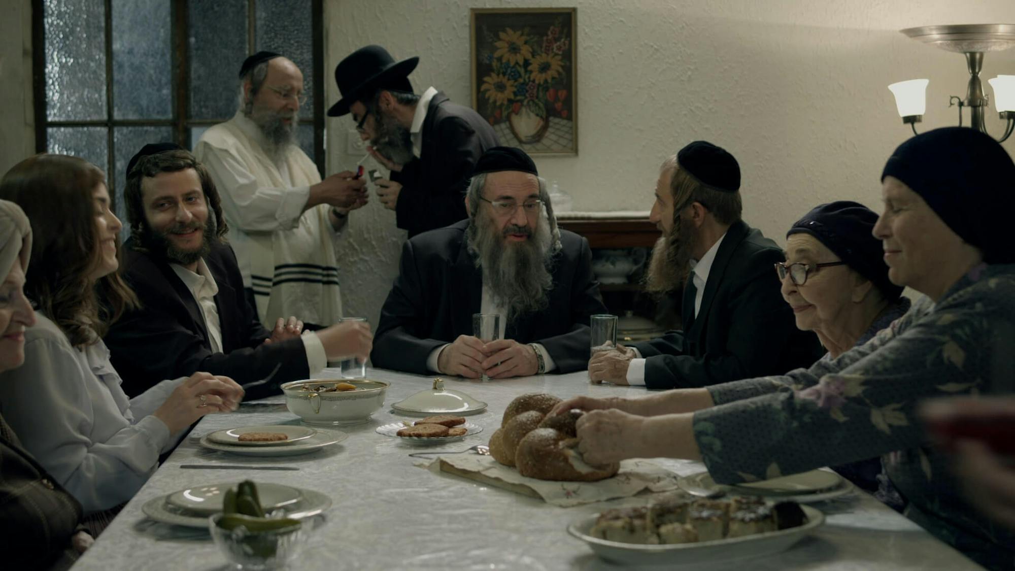 A shot from a Shtisel family meal. Shulem sits at the head, and the guests chat amongst themselves and break into the challah. The scene is lit by a small lamp.