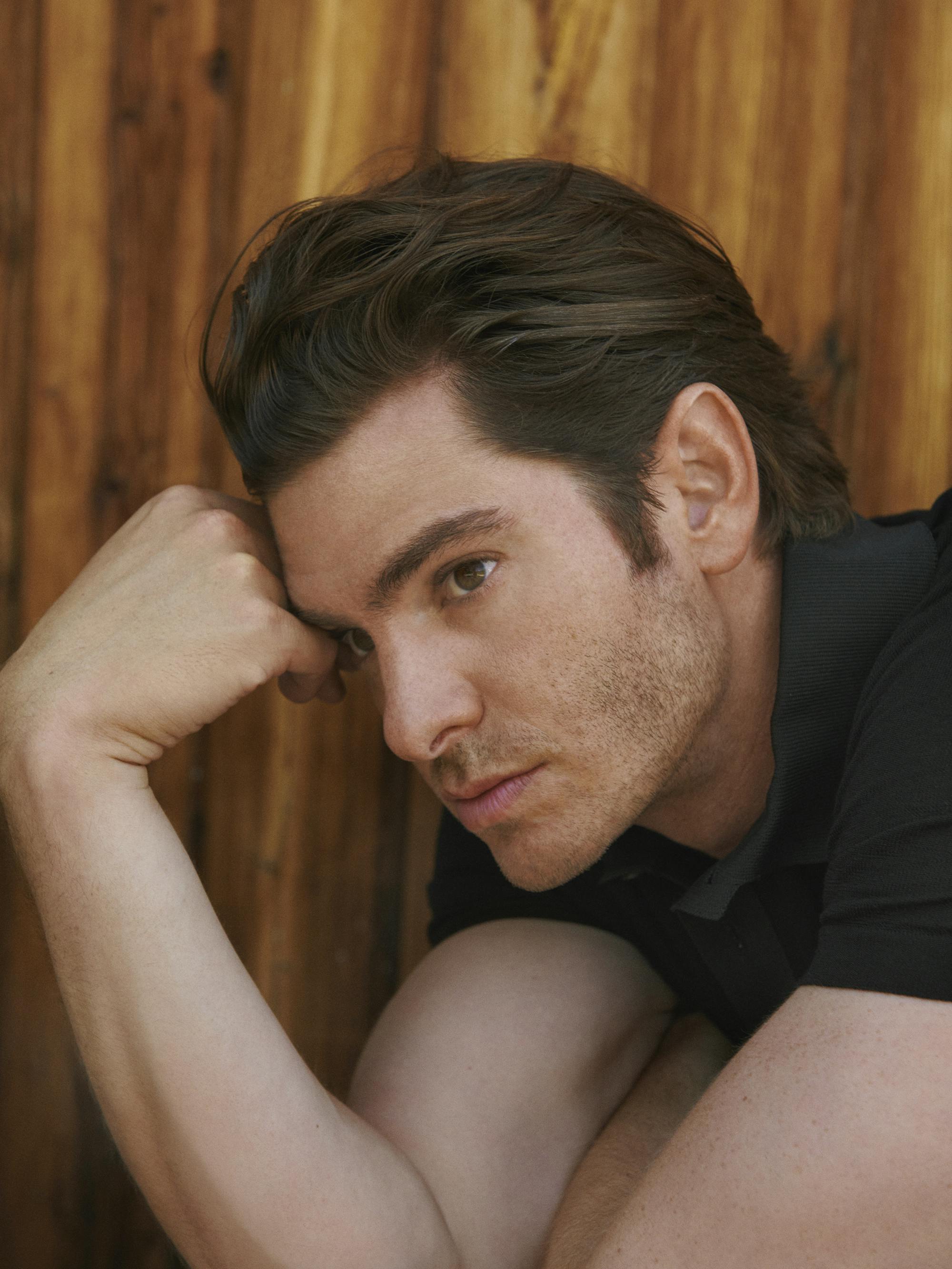 Andrew Garfield wears a black t-shirt and rests against a wooden wall.
