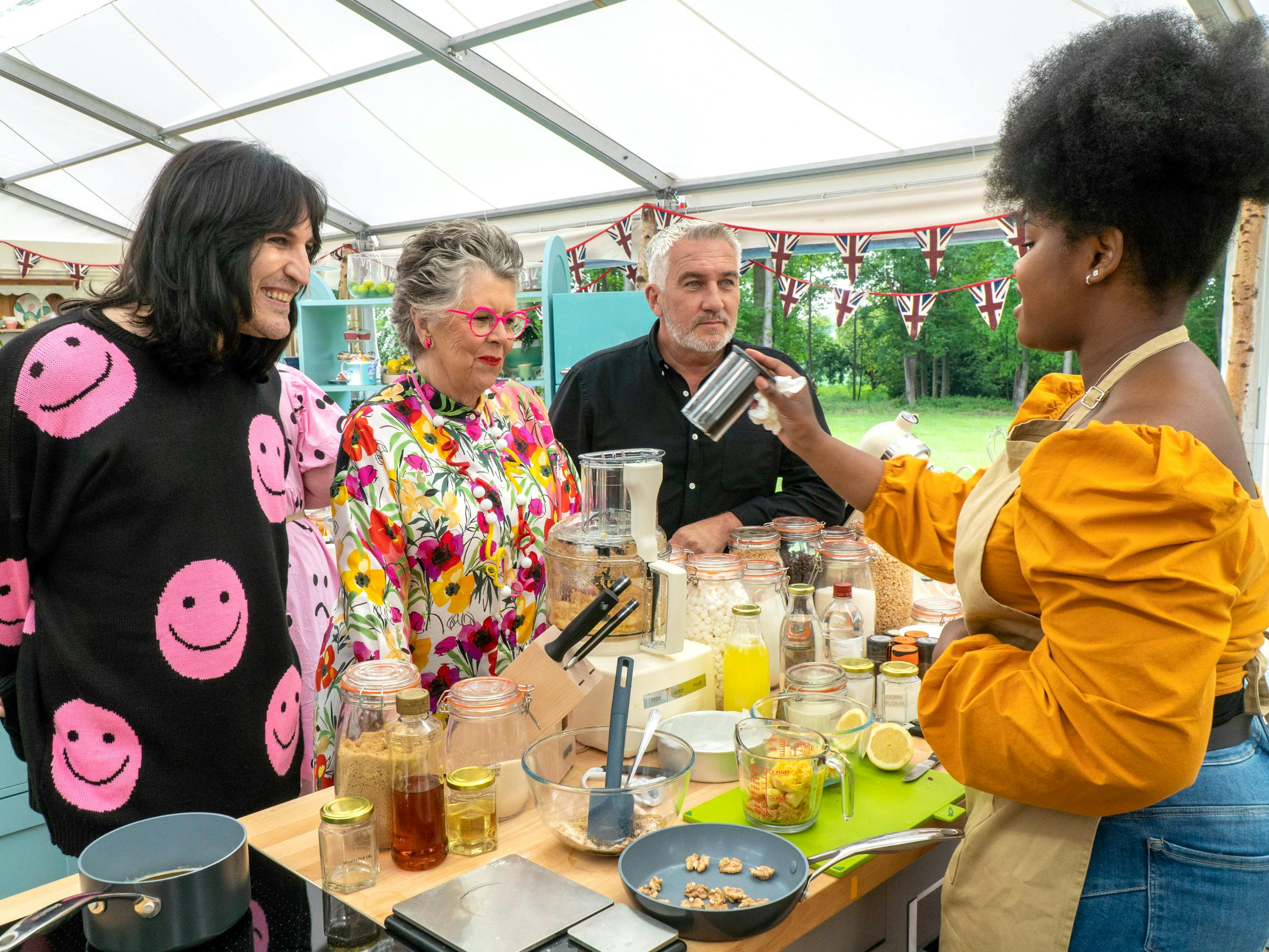 Noel Fielding, Mary Berry, Paul Hollywood, and contestant Rochica stand around a prep counter in the famed tent of The Great British Baking Show.