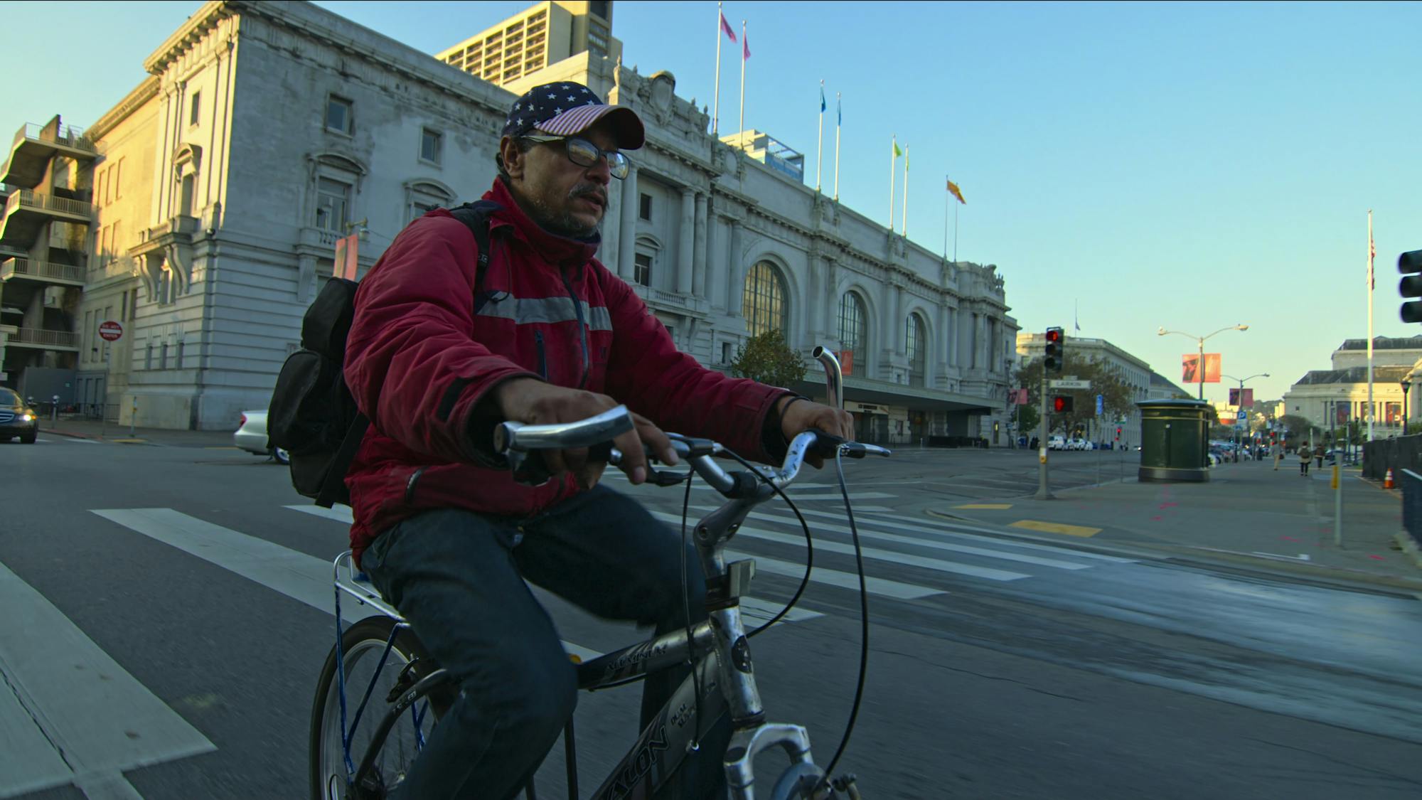 Luis Rivera Miranda wears grey pants and a red sweater. He bikes through a street with a old building behind him.
