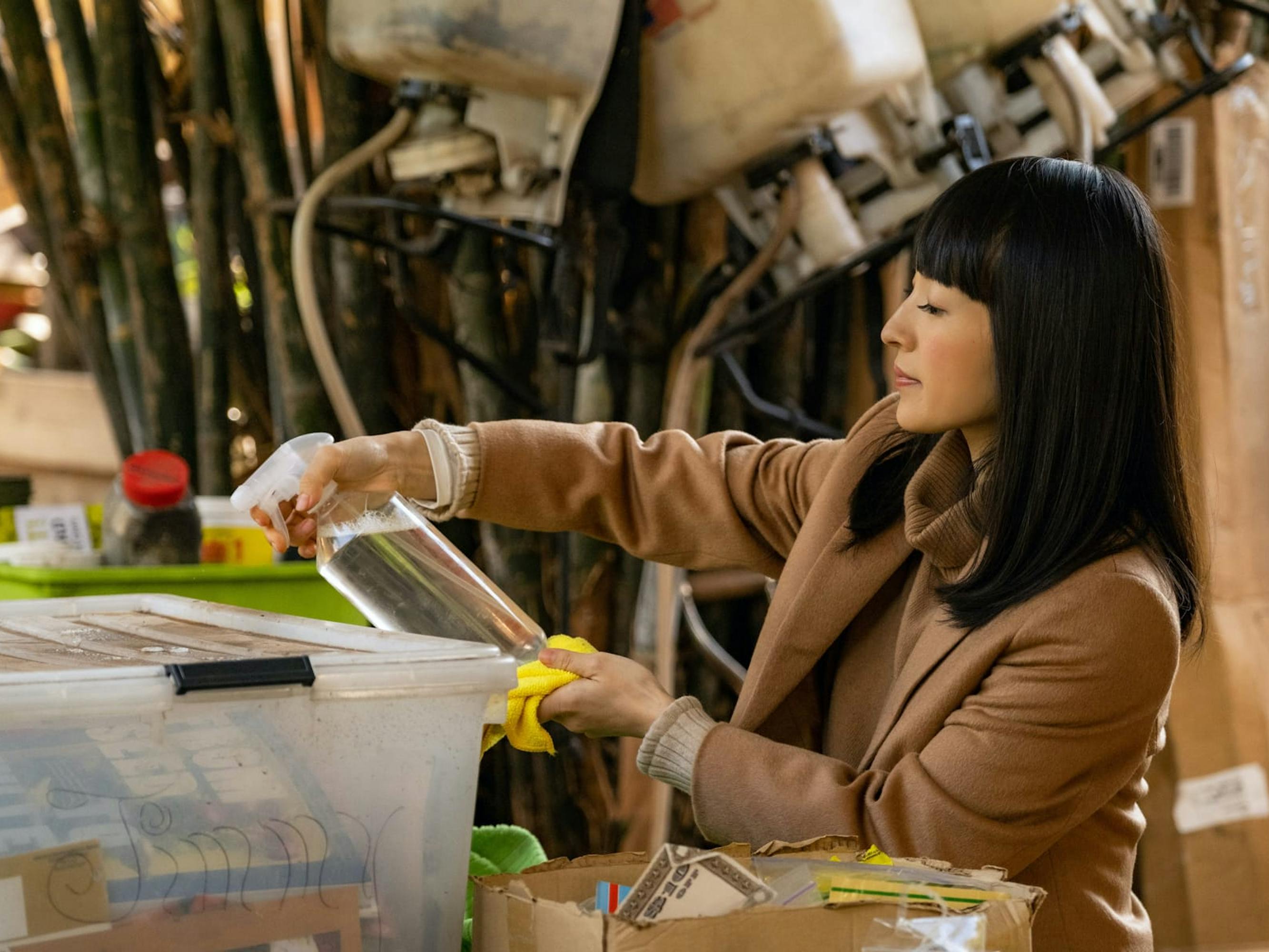 Marie Kondo uses a spray bottle to clean the top of a plastic storage bin coated in a thick layer of dust in a space filled with boxes and industrial equipment.
