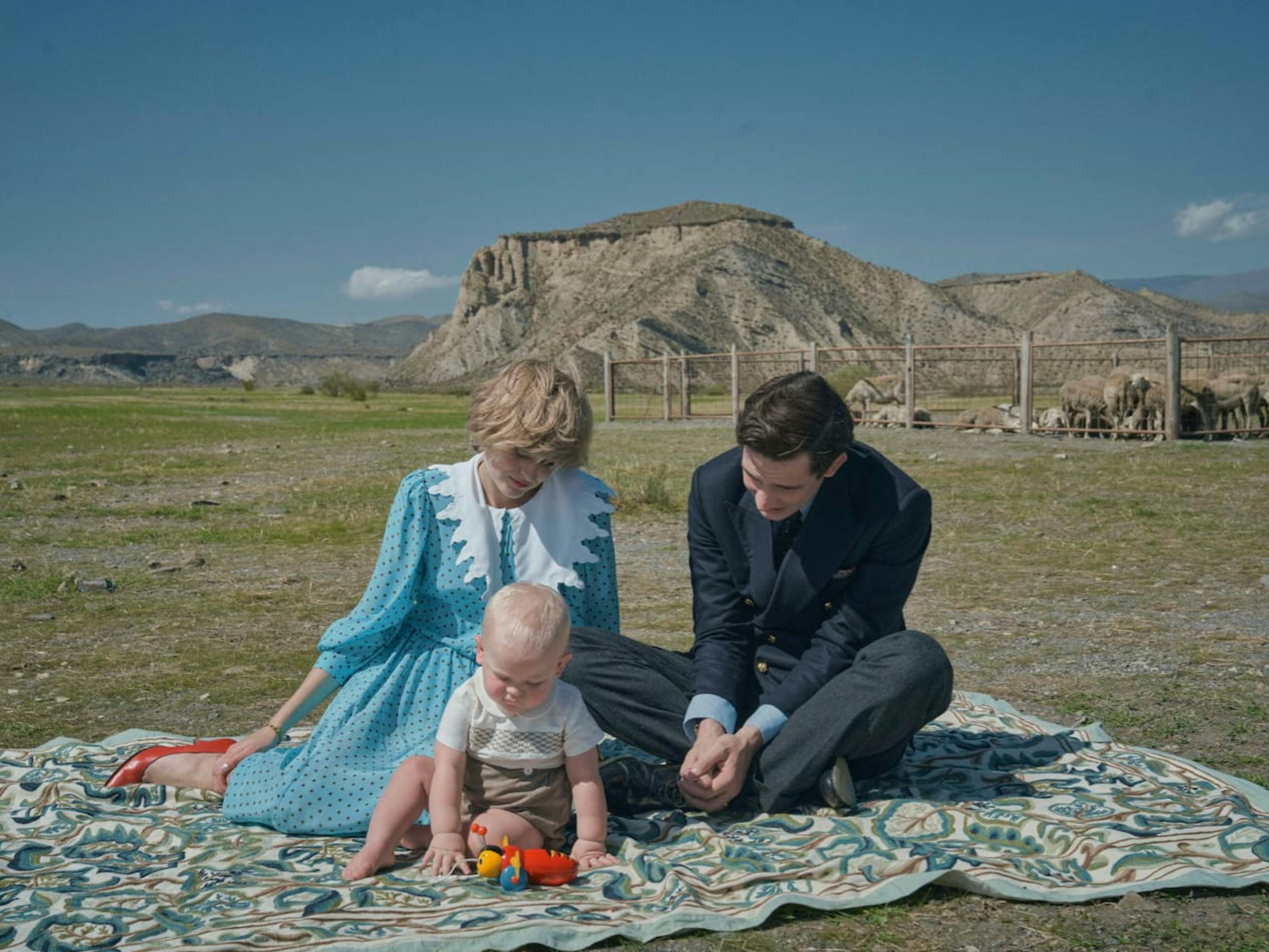 Princess Diana (Emma Corrin) and Prince Charles (Josh O’Connor) sit on a picnic blanket with their son. There is a blue sky and plateau behind them.