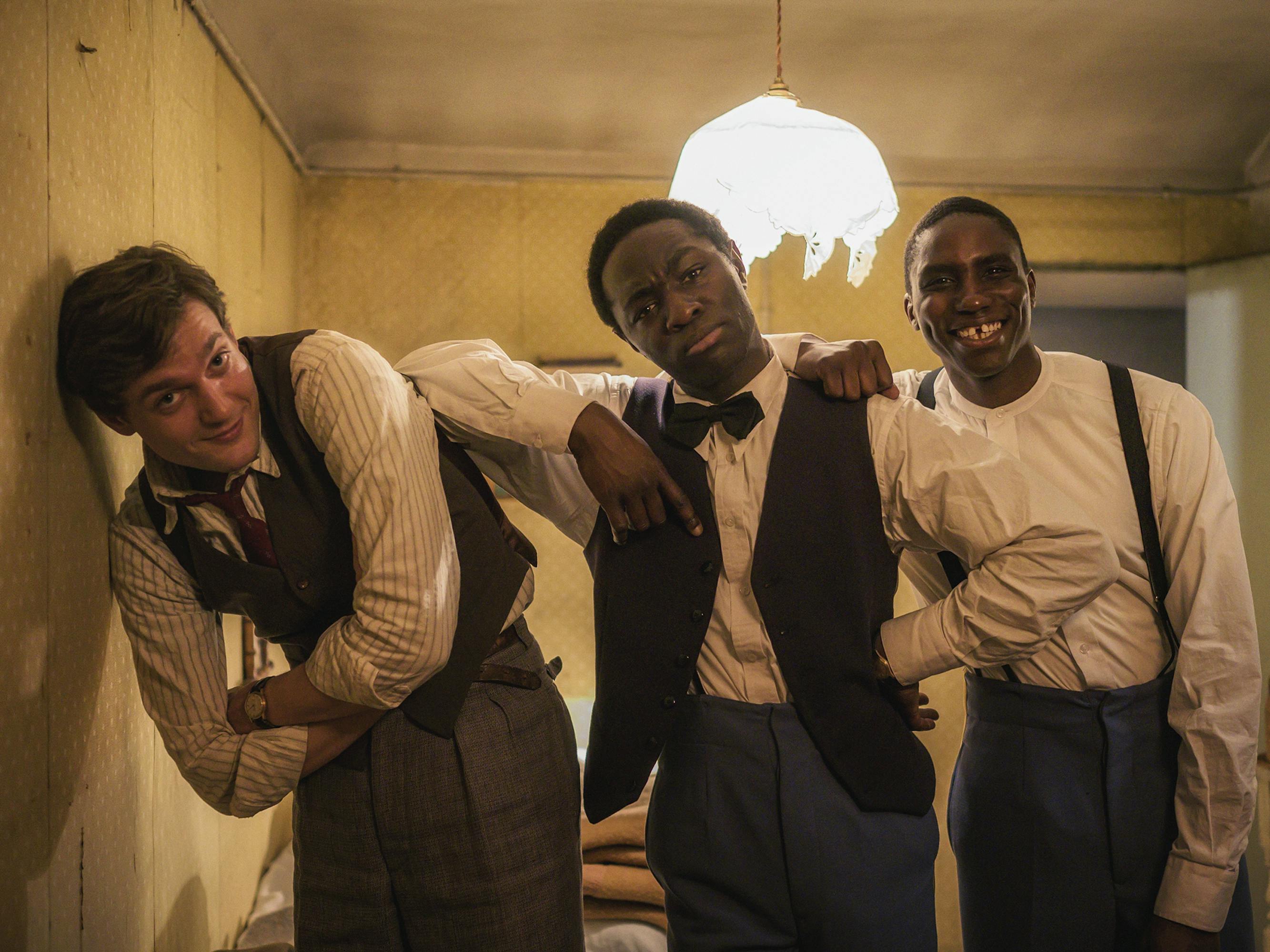 Albert Hirschman (Lucas Englander), Paul Diallo (Ralph Amoussou), and Jacques "Petit" Kandjo (Birane Ba) wear white shirts and dark dress pants, and lean against each other smiling — such a dapper crew!