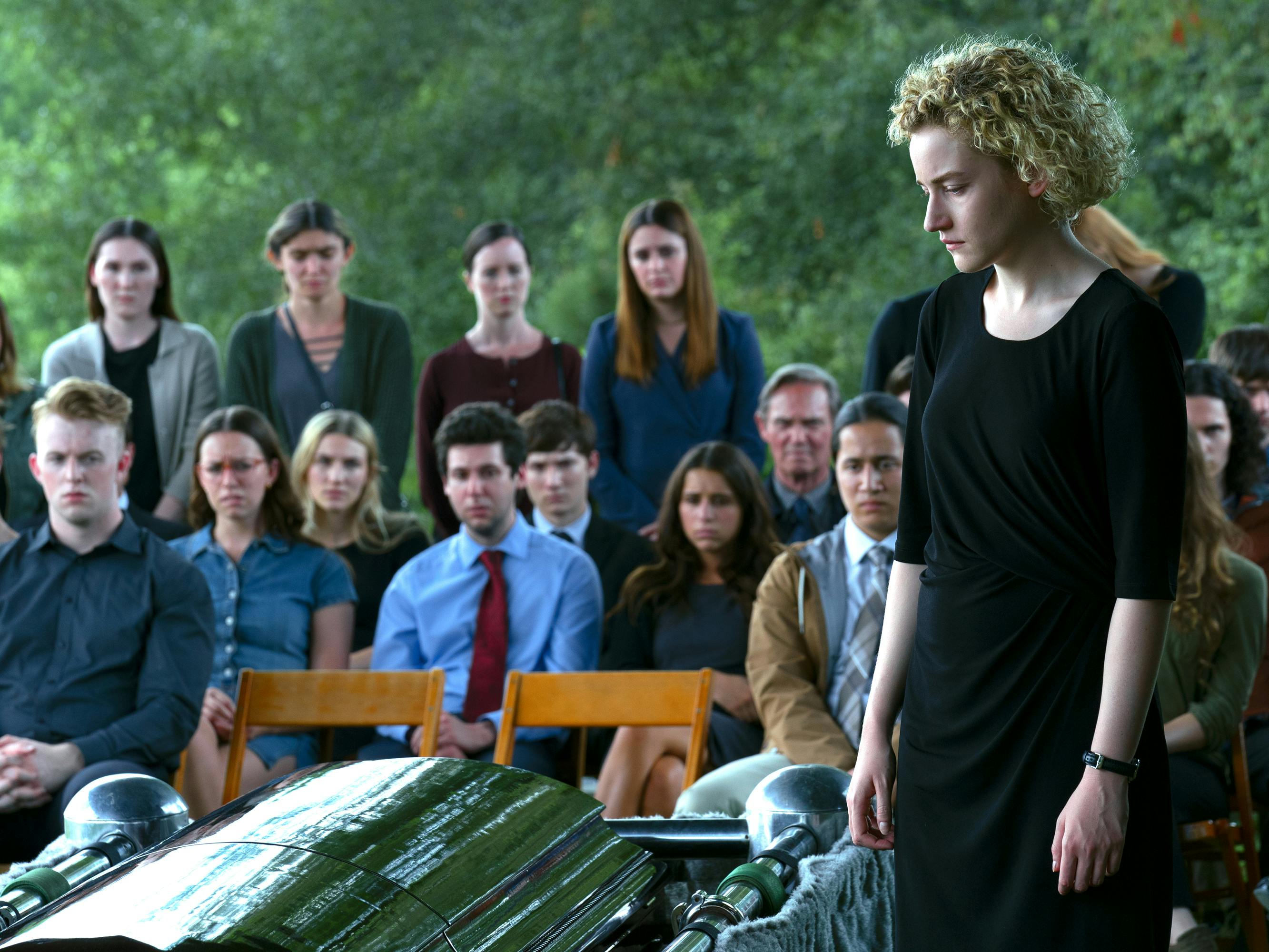 Ruth Langmore (Julia Garner) wears a black outfit and stands in front of a crowd of people. She looks down at a green coffin with a heartbroken look on her face. 