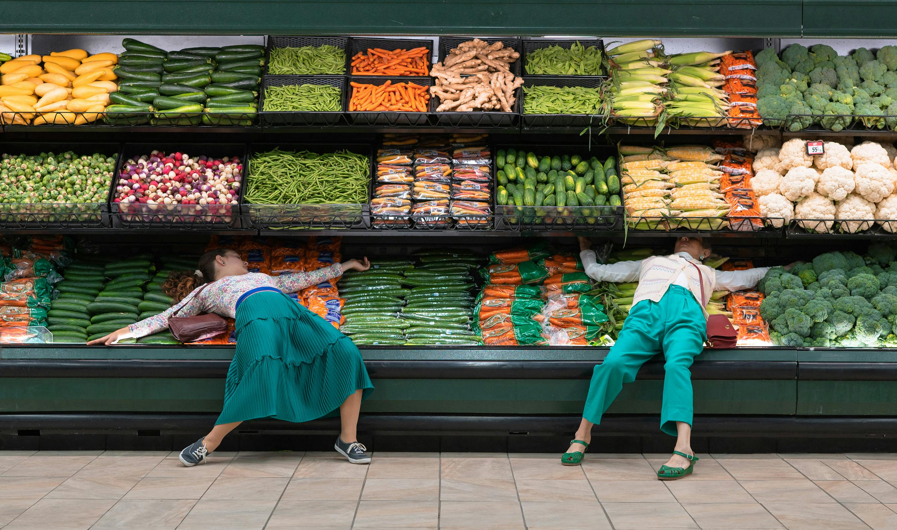 Two figures lie on the produce. They wear green pants and light tops.