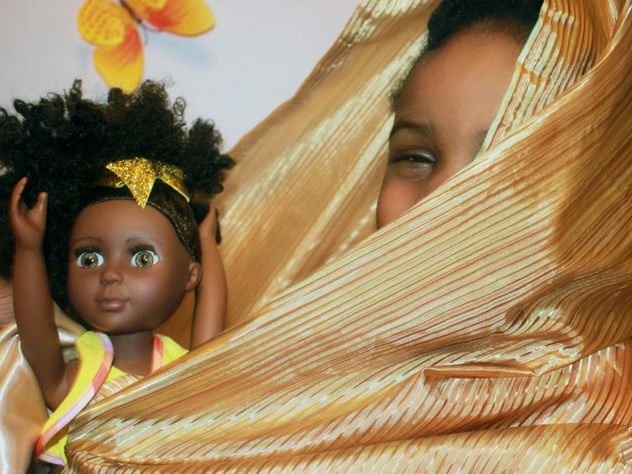 This photograph shows a girl with her doll. The girl is wrapped in gold, striped fabric that shines in the artificial light. Only part of her face is visible. Her doll is posed with its hands thrown upwards. The image of an orange butterfly is visible in the background.