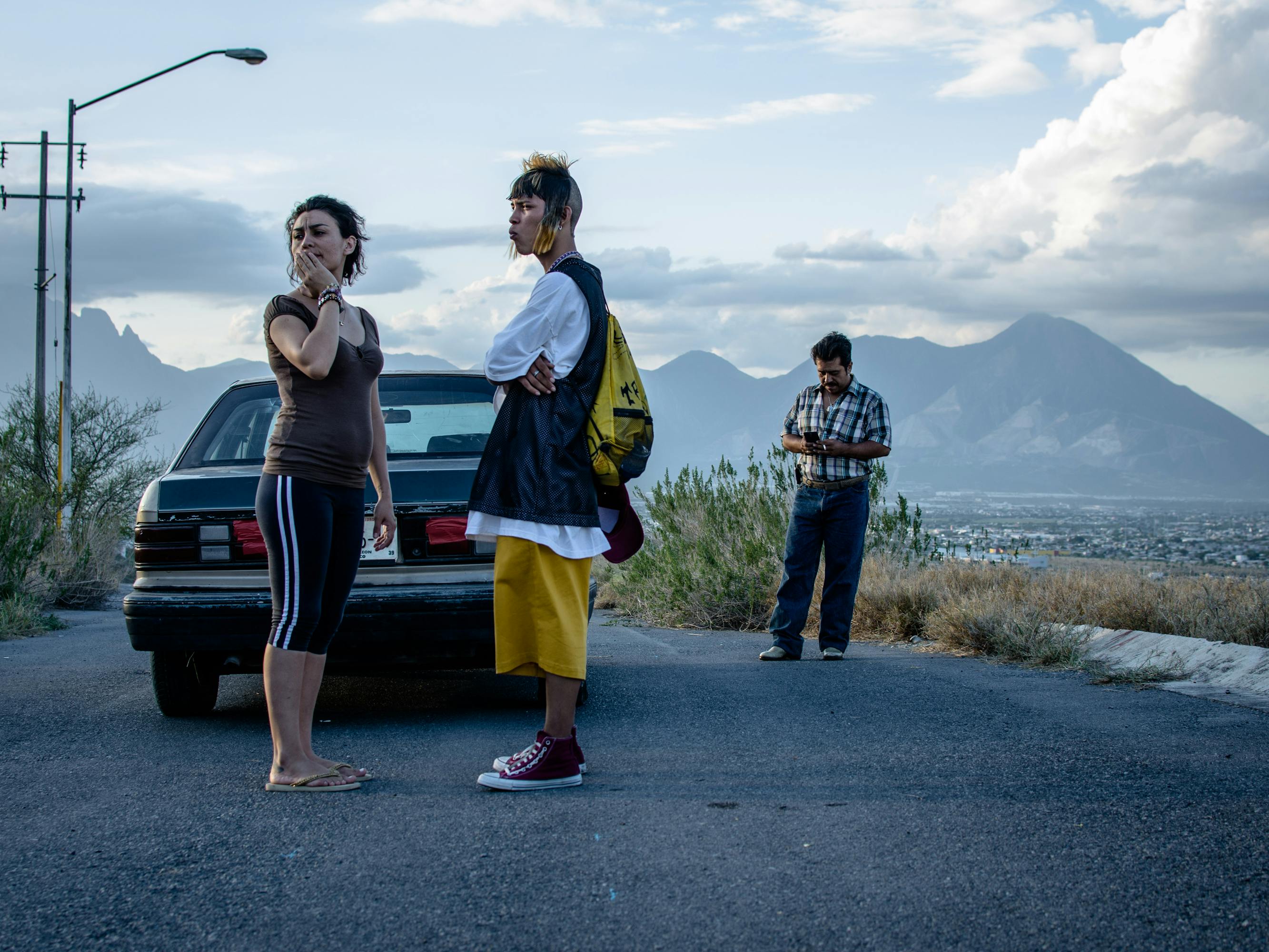 Three characters from Ya no estoy aquí stand against a mountain range by a black car.