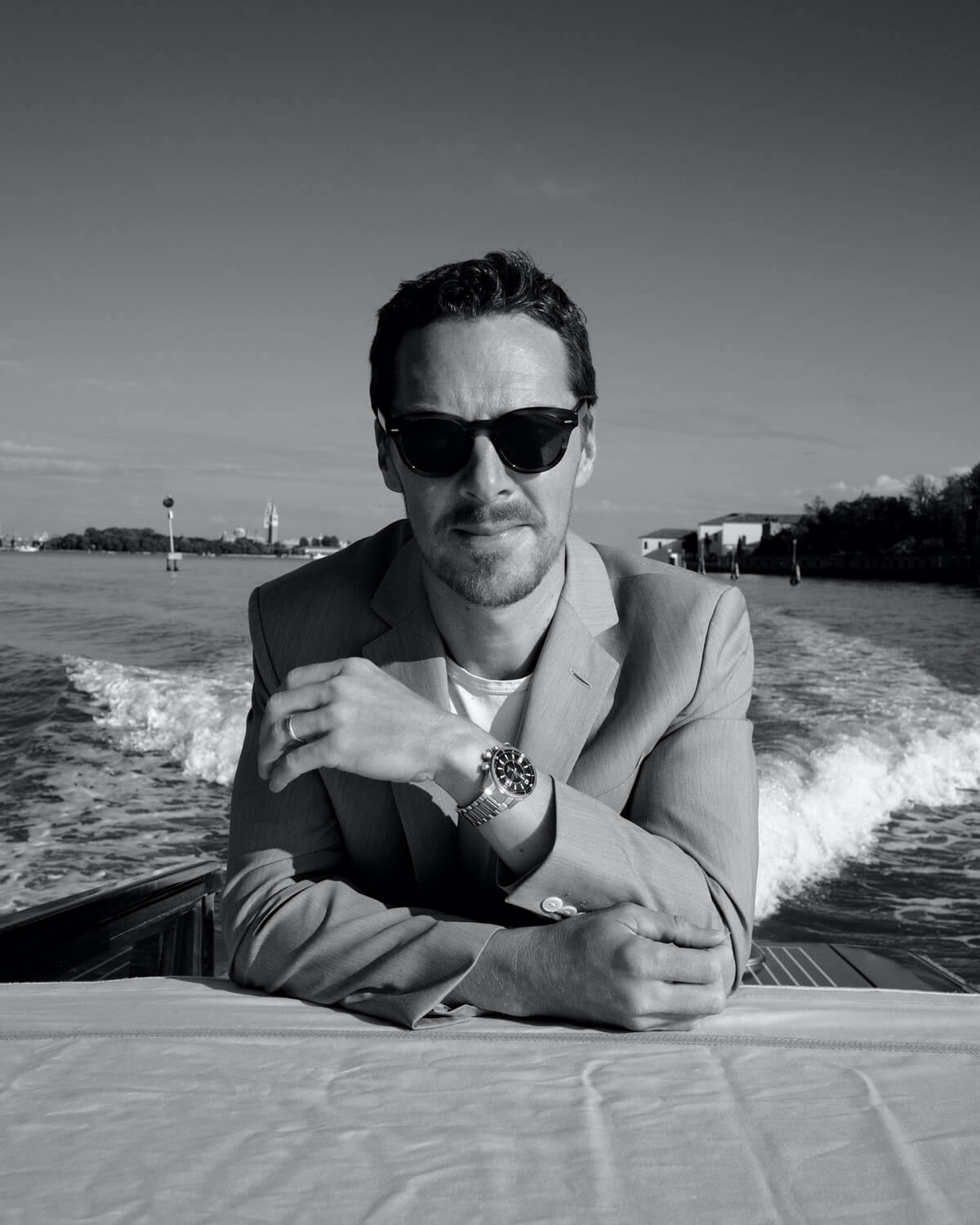 Benedict Cumberbatch wears a light suit, dark glasses, and a watch. He poses in the middle of a boat, speeding around Venice.