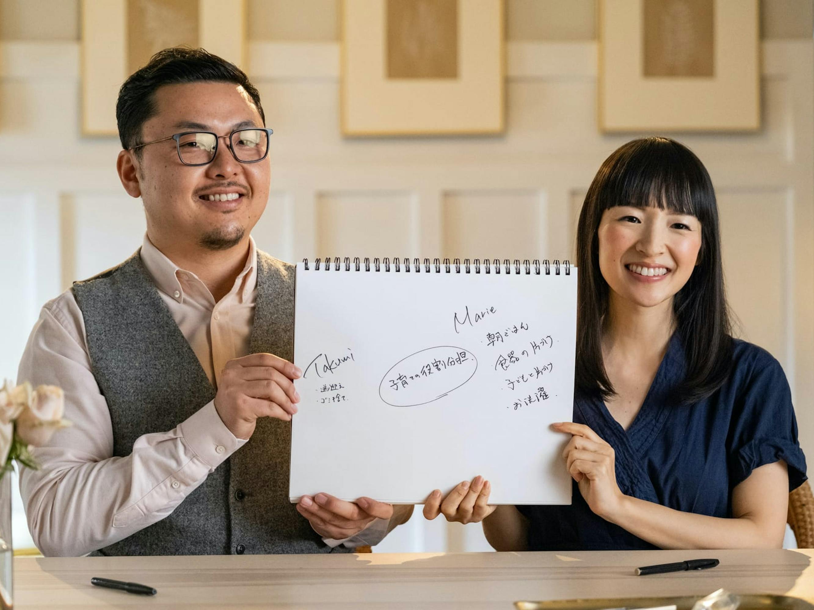 Marie Kondo’s husband Takumi and Marie Kondo sit at a table and hold up a sketchpad between them with their names and some writing in kanji. On the table in front of them are pens and a vase of light pink roses.