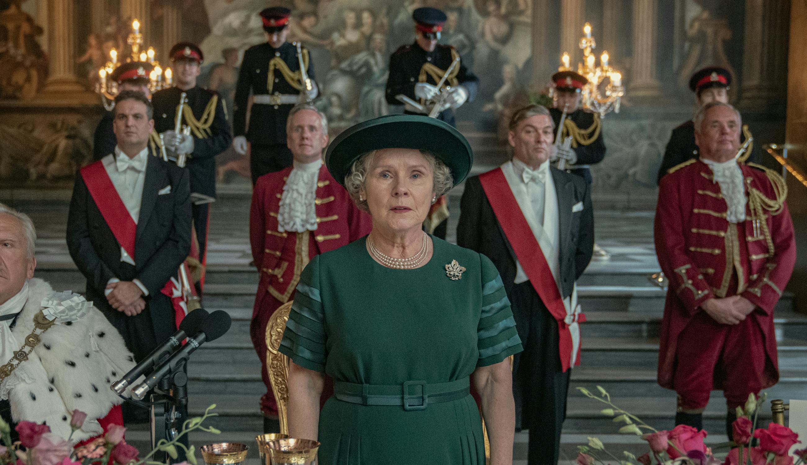 Queen Elizabeth (Imelda Staunton) wears green and stands in front of some guards wearing white, black, and red.