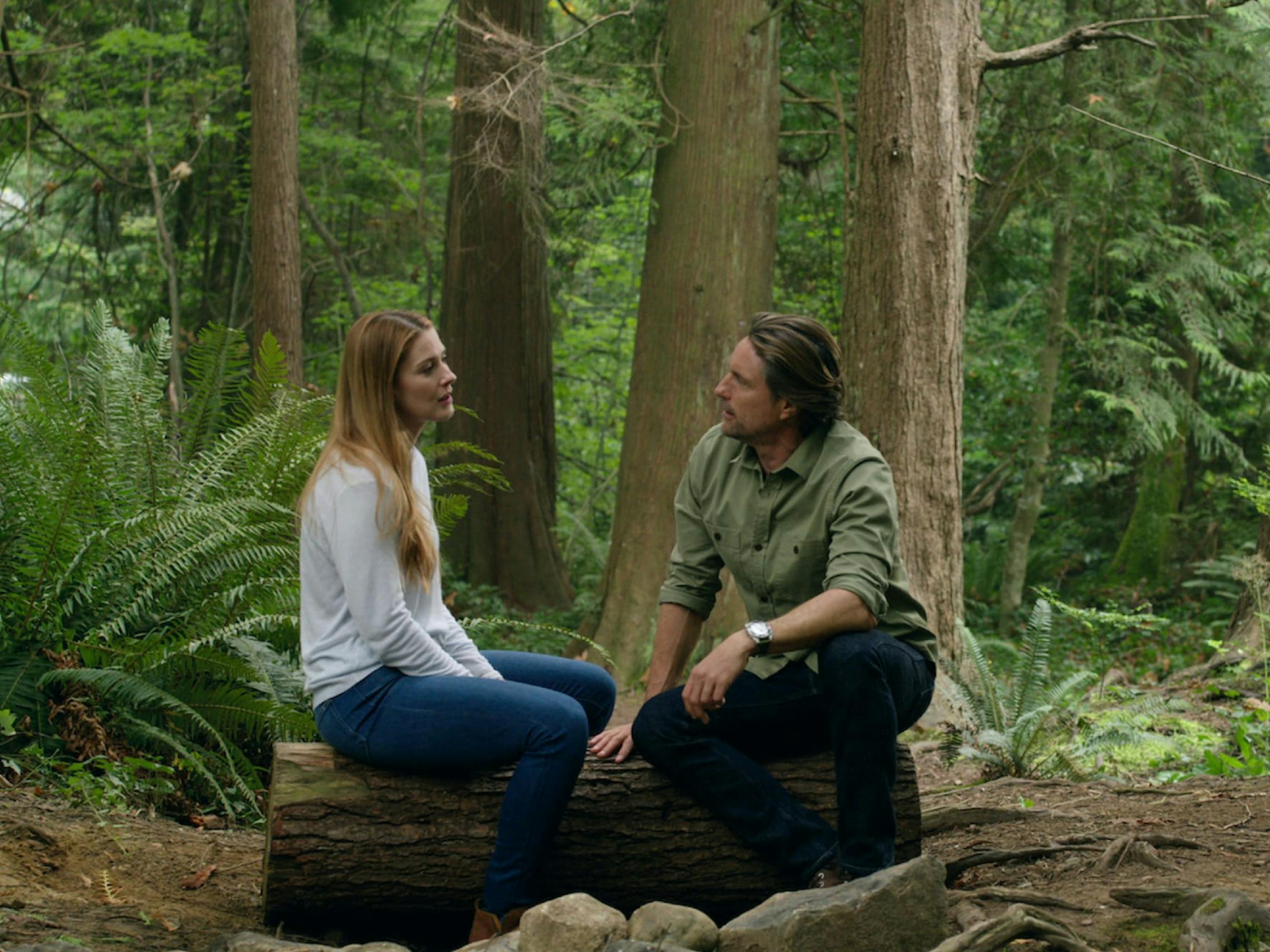 Melinda Monroe (Alexandra Breckenridge) and Jack Sheridan (Martin Henderson) sit on a log in the middle of a forest. She wears a white shirt and jeans, and he wears a green shirt and jeans.
