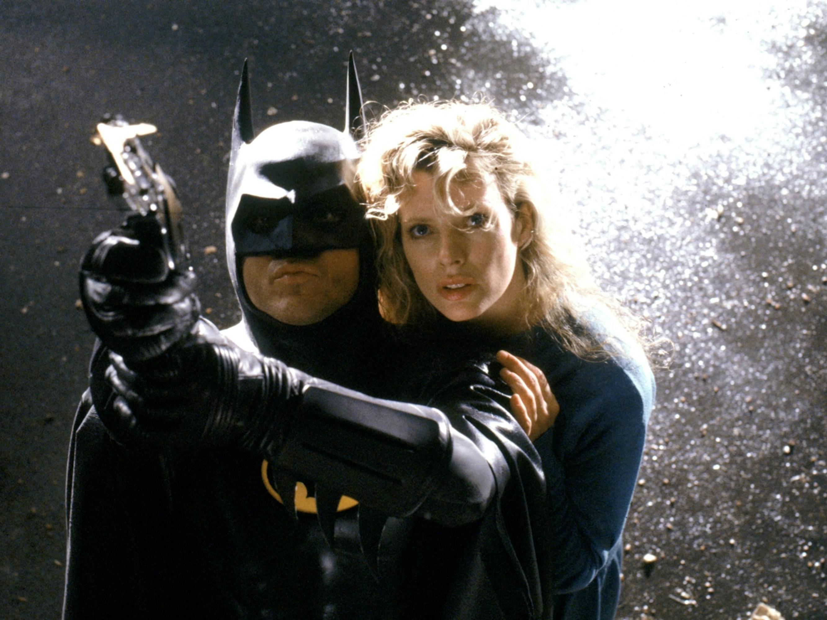 Batman (Michael Keaton) and Vicki Vale (Kim Basinger) in Batman stand in a sparsely lit scene. Vicki hovers behind a suited Batman holding a gun.