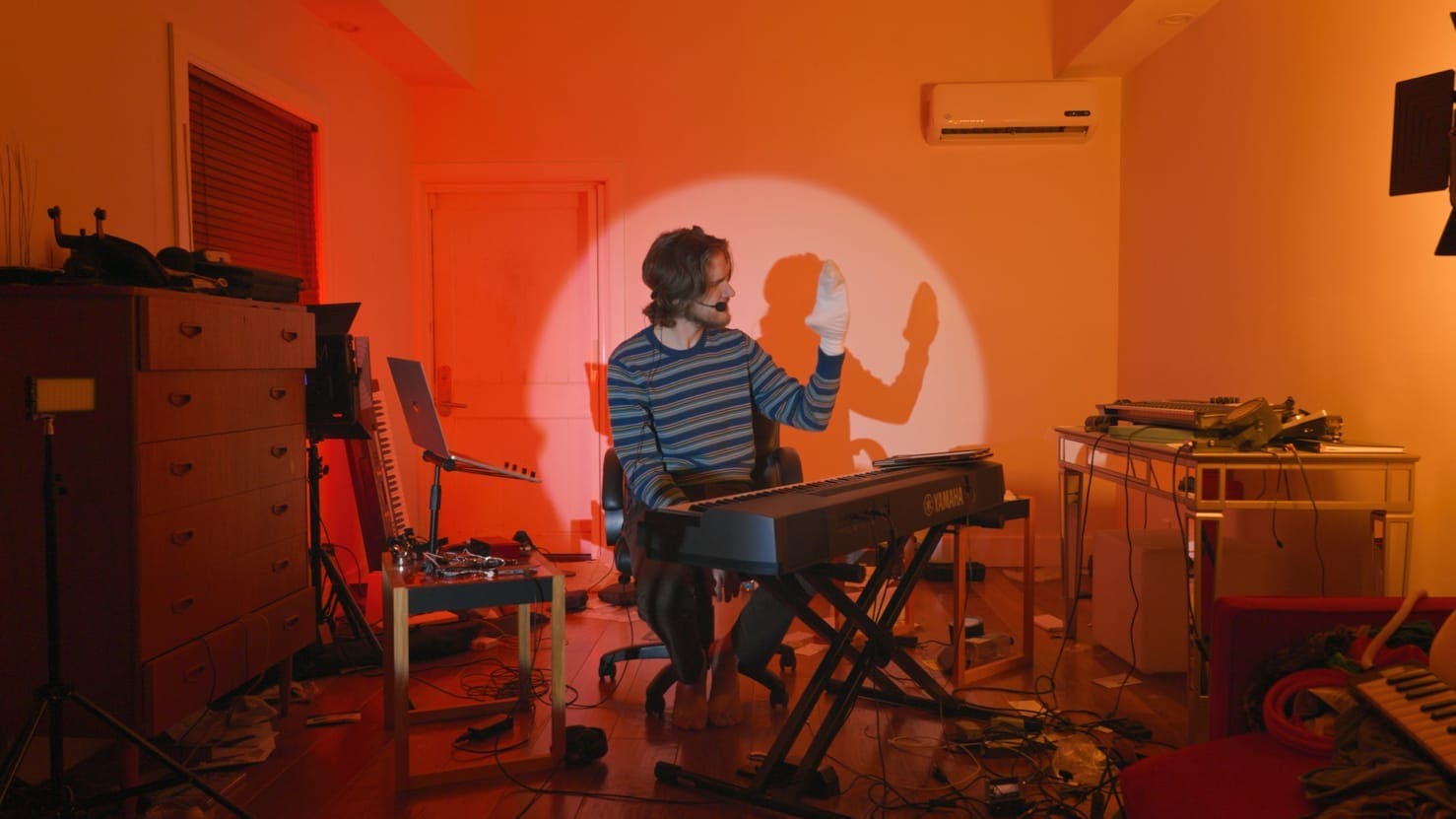 Bo Burnham performs on a keyboard with a sock puppet. He is lit by a spotlight, and the room is tinted red, orange, and yellow. There is also a dresser, computer and stand, table, and stool in the room. The floor is messy with a range of objects, mainly cords.