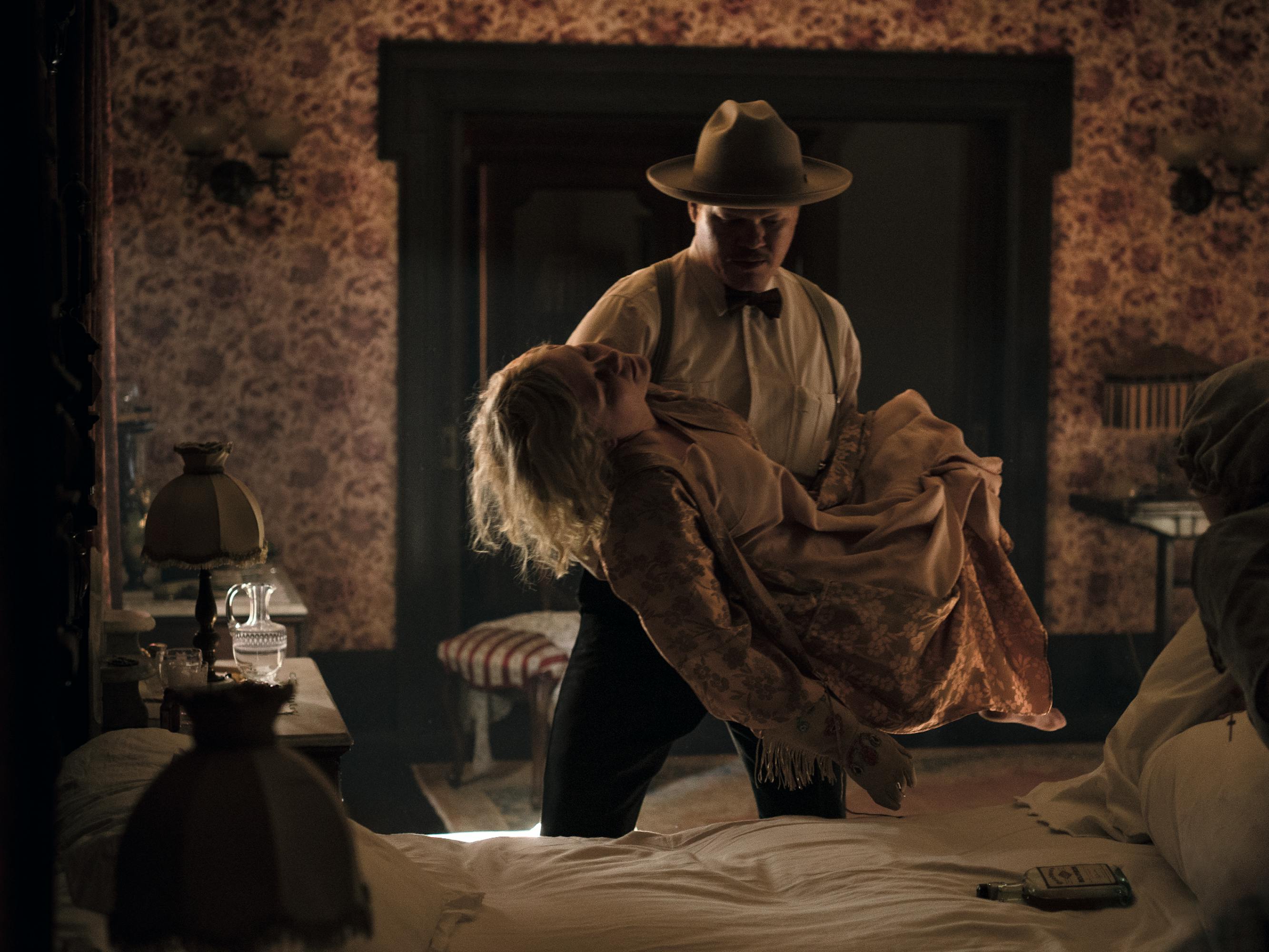 Jesse Plemons carries Kirsten Dunst's limp body to her bed. He wears a white shirt, black bowtie and wide-brimmed hat. Dunst wears a pink dress.