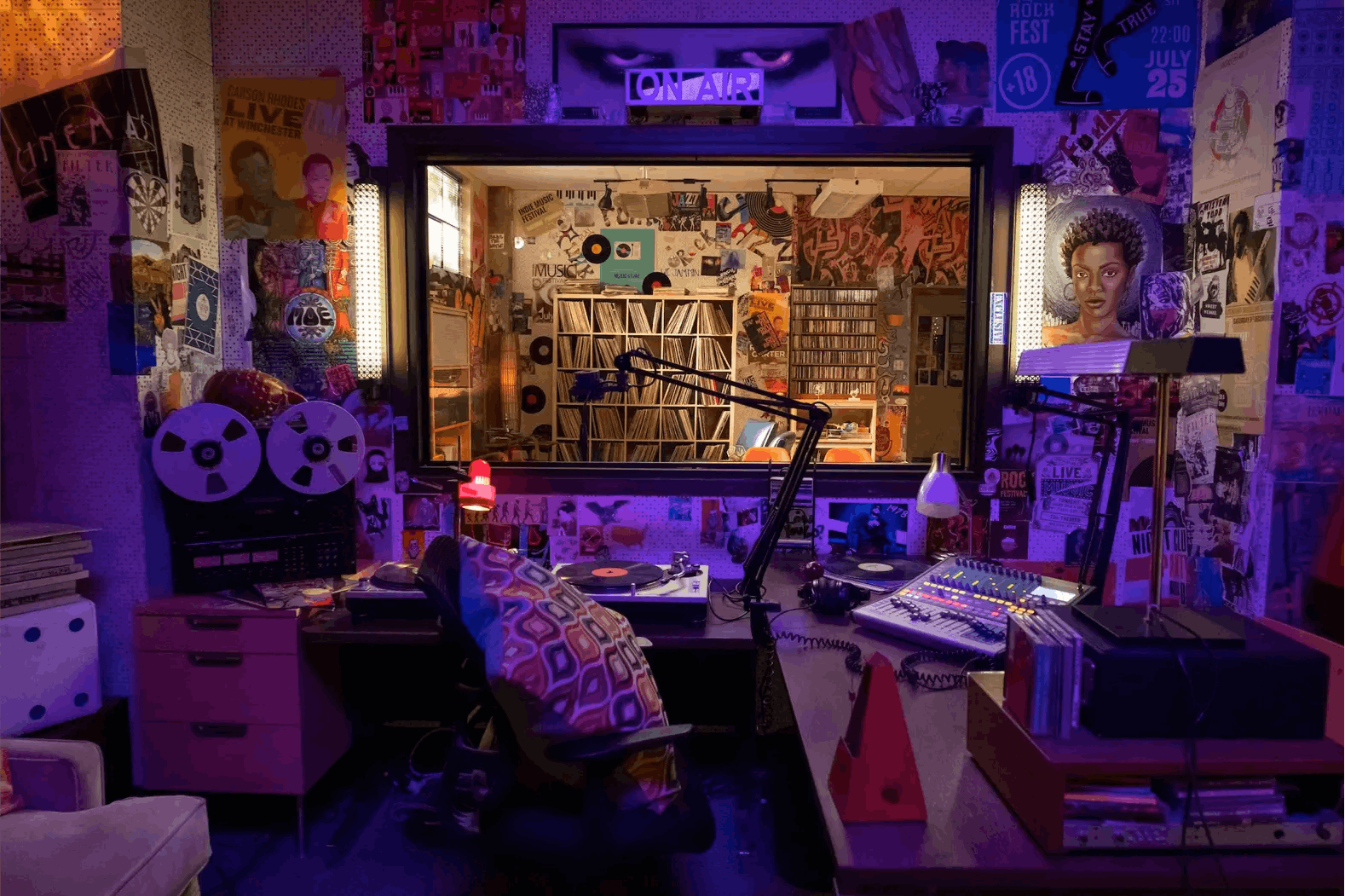A blue lit shot of the inside of the recording booth. The walls are lined with art and posters and there is a massive mic in the middle of the room.