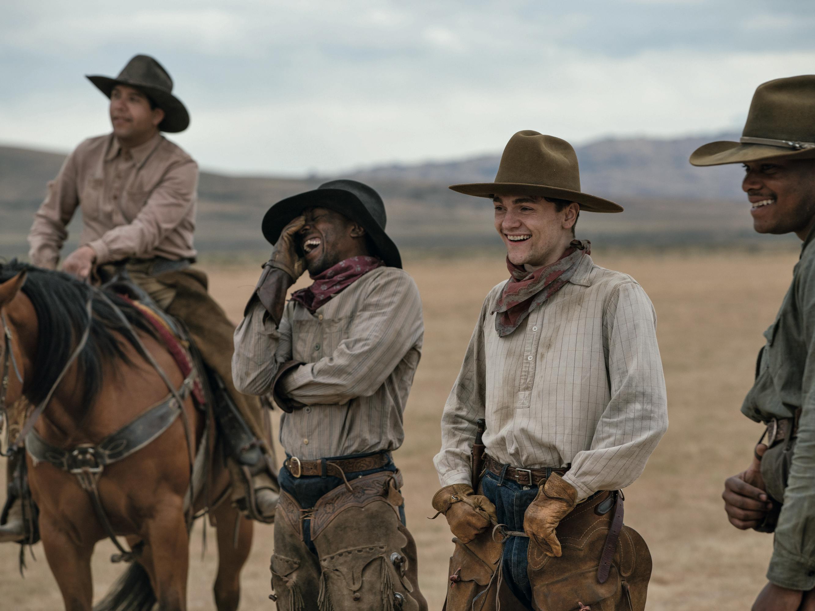Ranch hands from The Power of the Dog's Burbank family stand laughing in front of a mountain range in the distance.