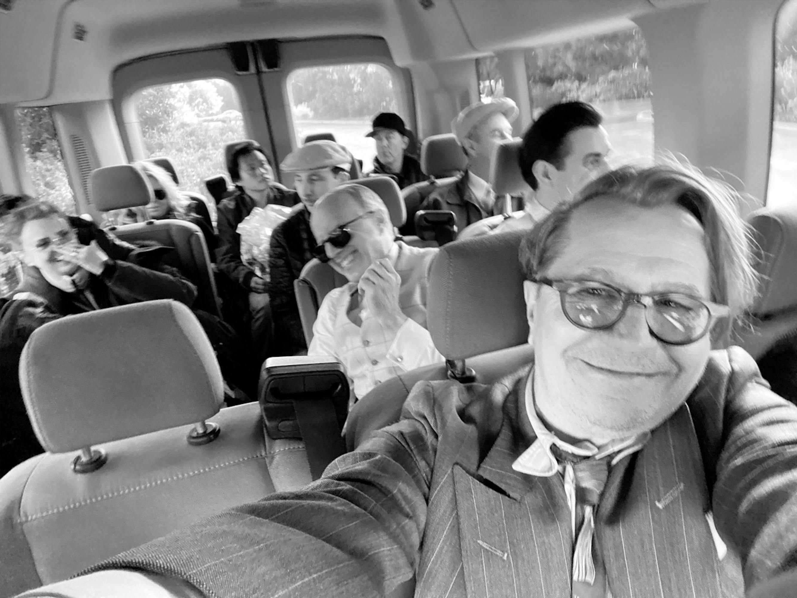 Oldman takes a smiling selfie in a van, with cast and crew enjoying themselves in the background.