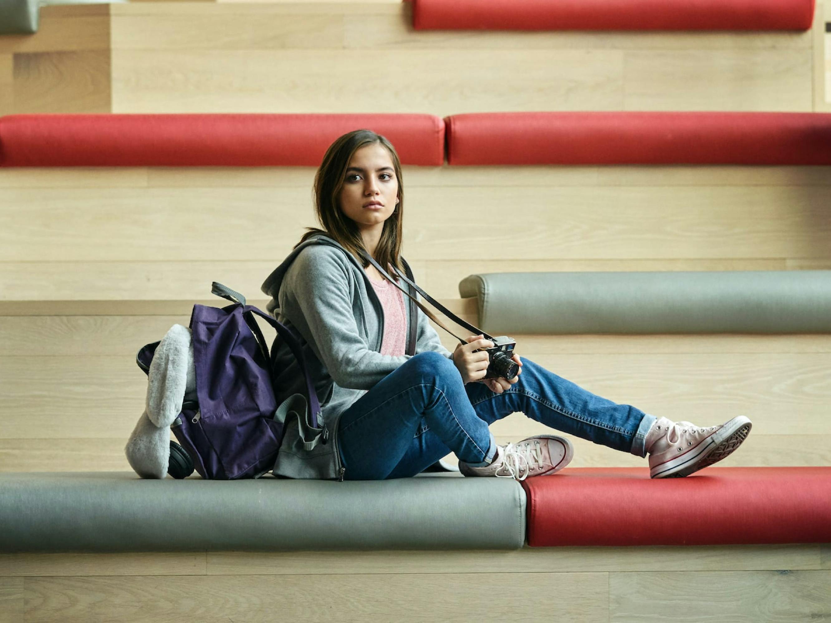 Rachel Cooper (Isabela Merced) in Sweet Girl. She sits on some red and grey bleachers with jeans and a purple backpack.