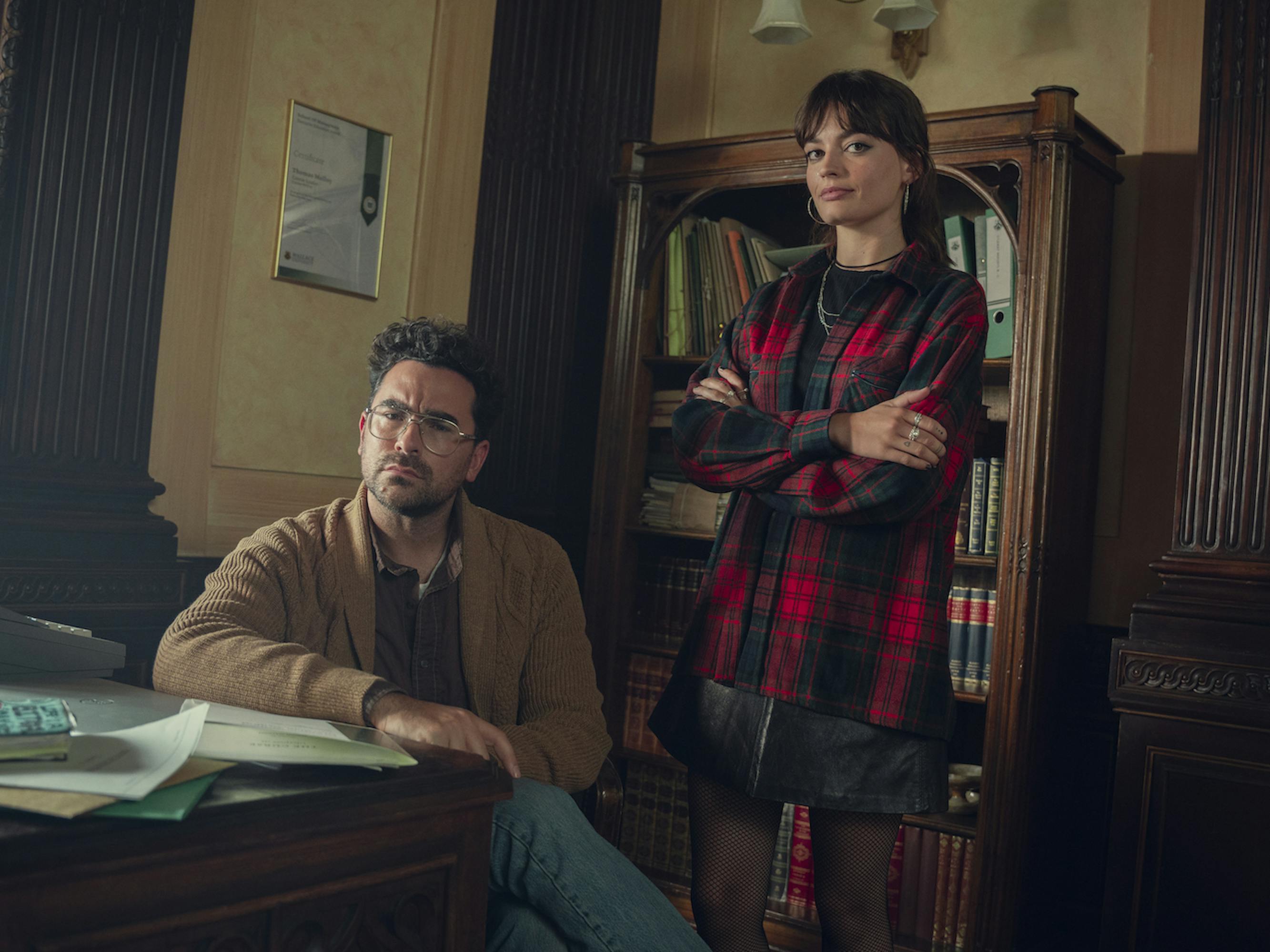 Thomas Molloy (Dan Levy) and Maeve Wiley (Emma Mackey) stand together in a wood paneled office with an American flag.