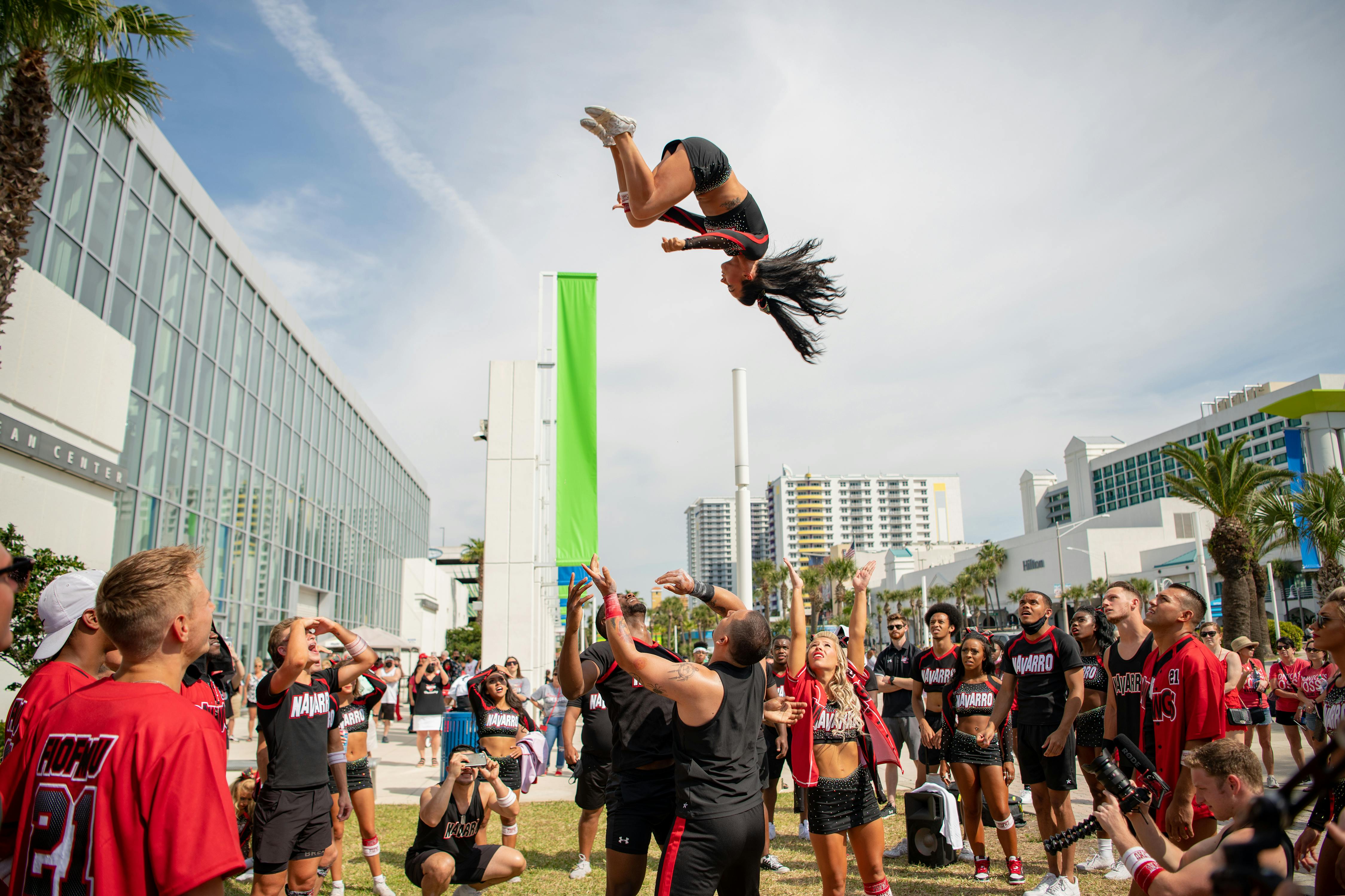 The Navarro cheer team circle up and throw a girl into the air. Behind them are expansive high rise buildings and a cloud-streaked sky.