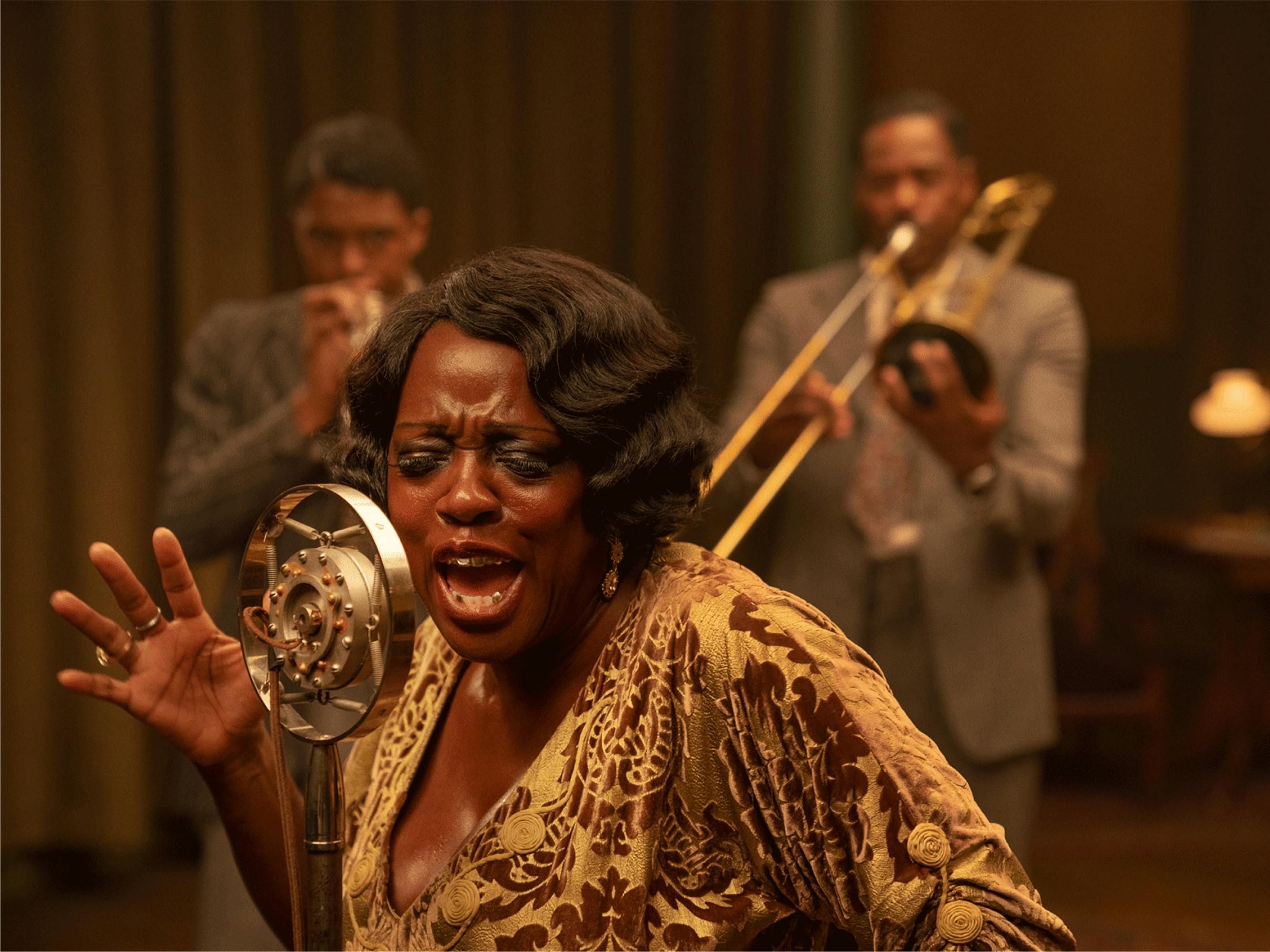 Ma Rainey (Davis) sings into her mic, one hand raised expressively. In the background, band members Levee (Boseman) and Cutler (Colman Domingo) play their horns.