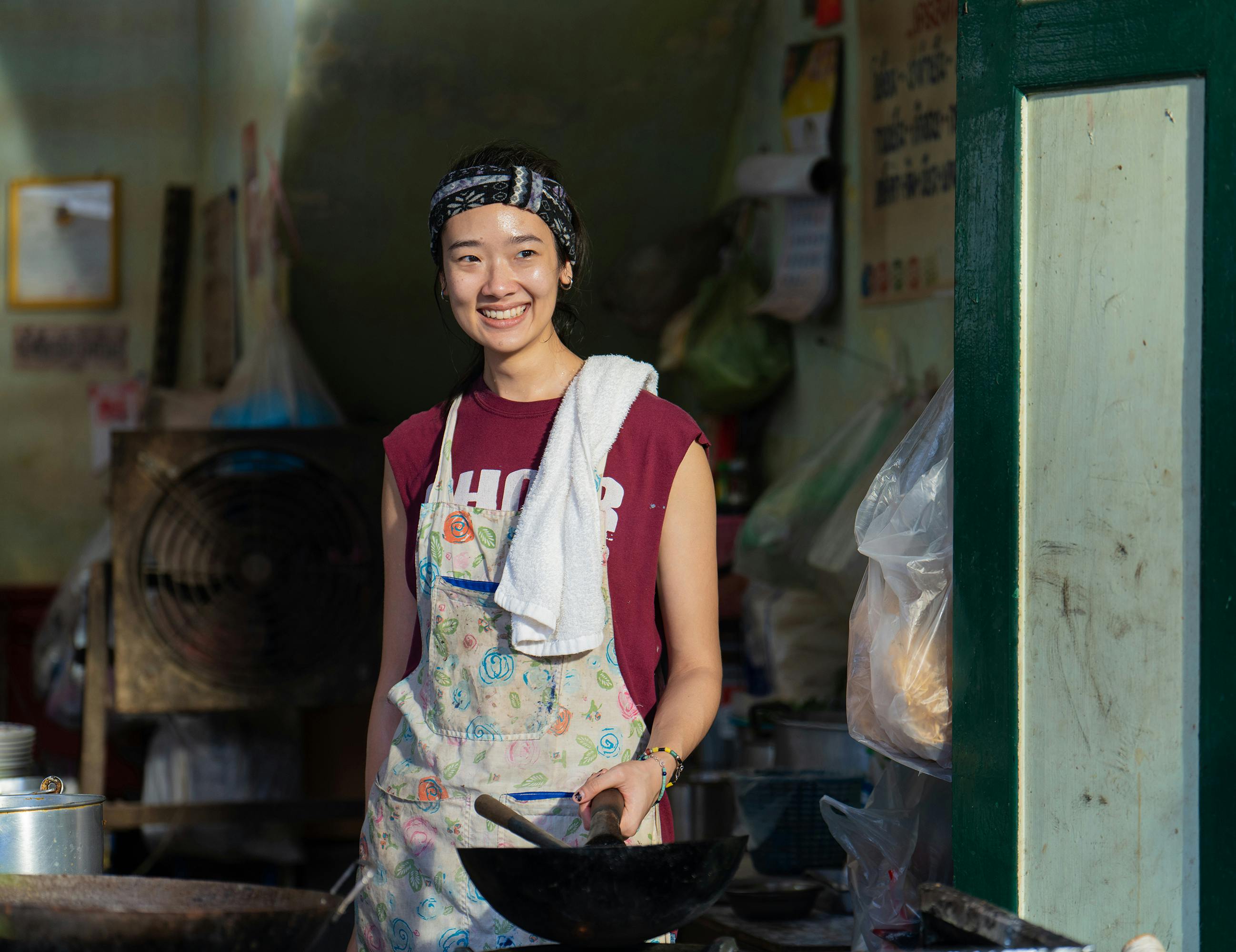 Aoy (Chutimon “Aokbab” Chuengcharoensukying) wears a red shirt and colorful apron and holds a wok, smiling at something offcamera