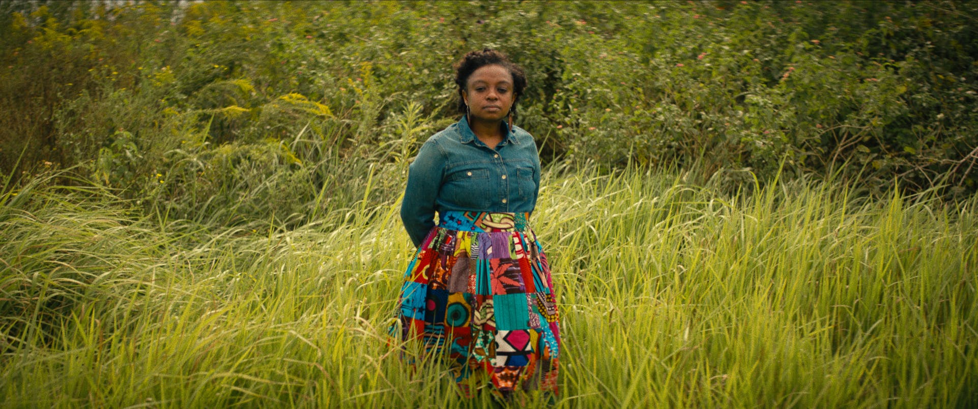Joycelyn Davis wears a denim long-sleeved shirt and patterned skirt, and stands in a field of tall grass.