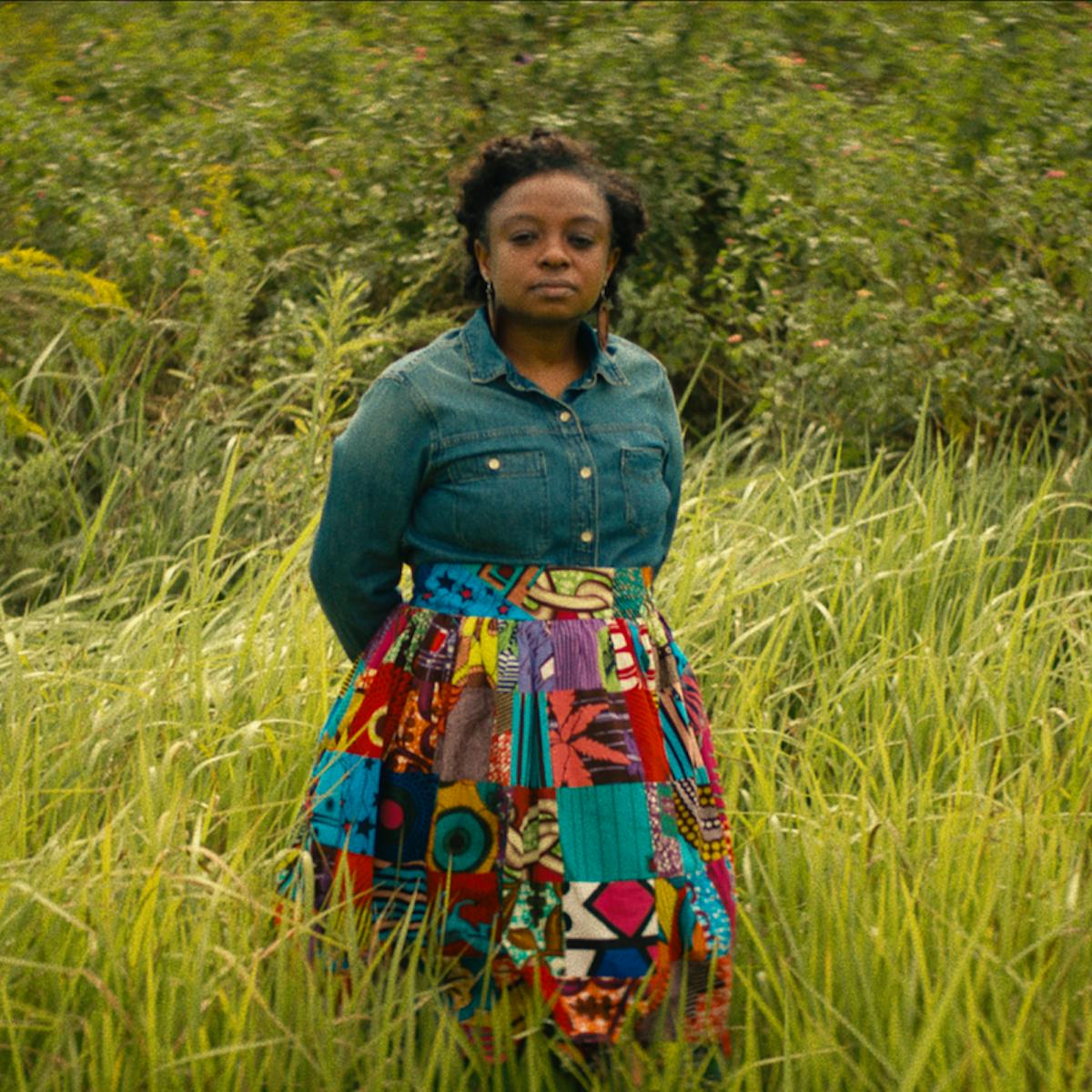 Joycelyn Davis wears a denim long-sleeved shirt and patterned skirt and stands in a field of tall grass.