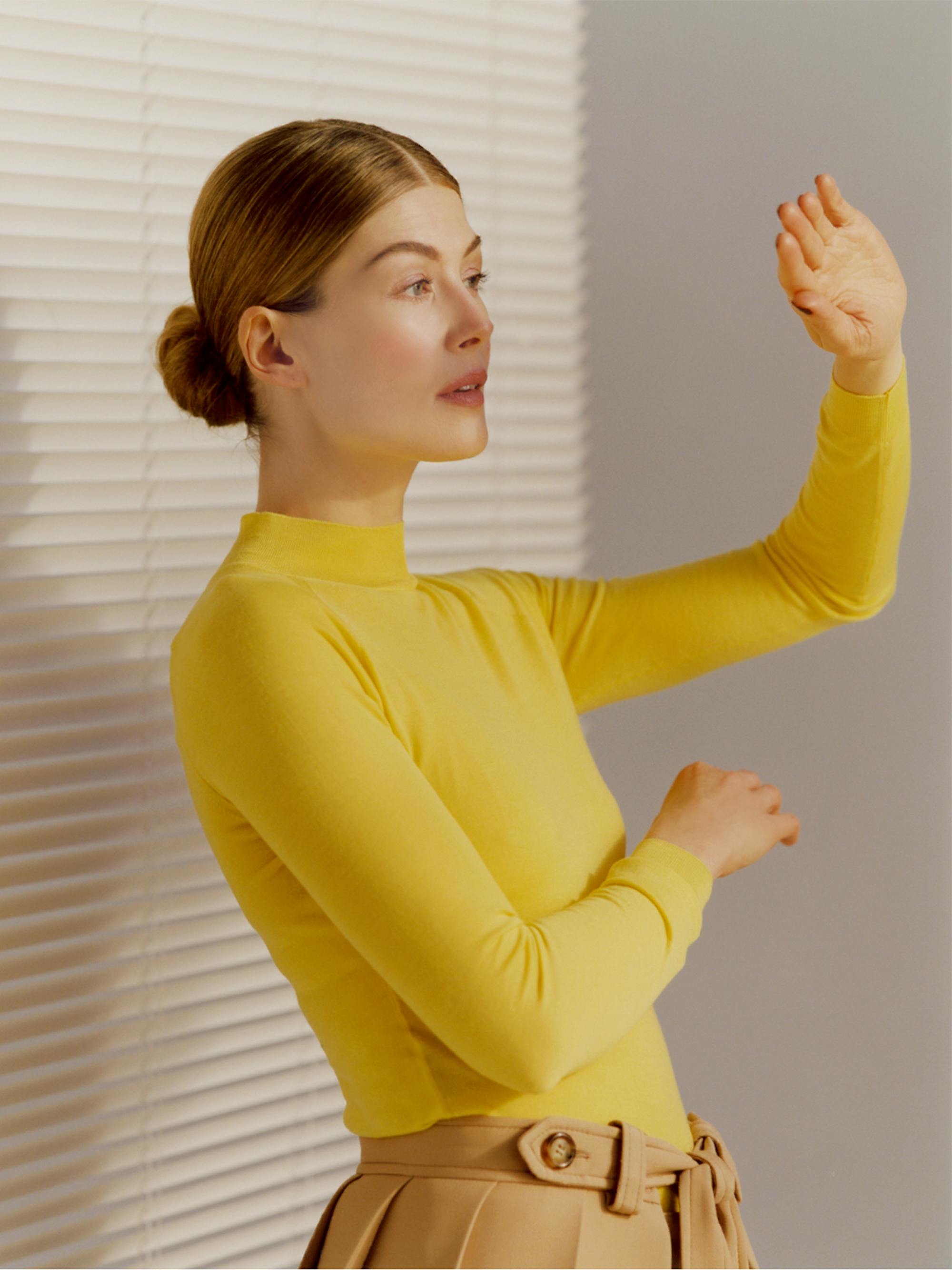 Rosamund Pike, posed in front of closed venetian blinds, wearing a bright yellow fitted turtleneck and beige trousers. She holds a hand up against the light pouring in from an unseen source in front of her.