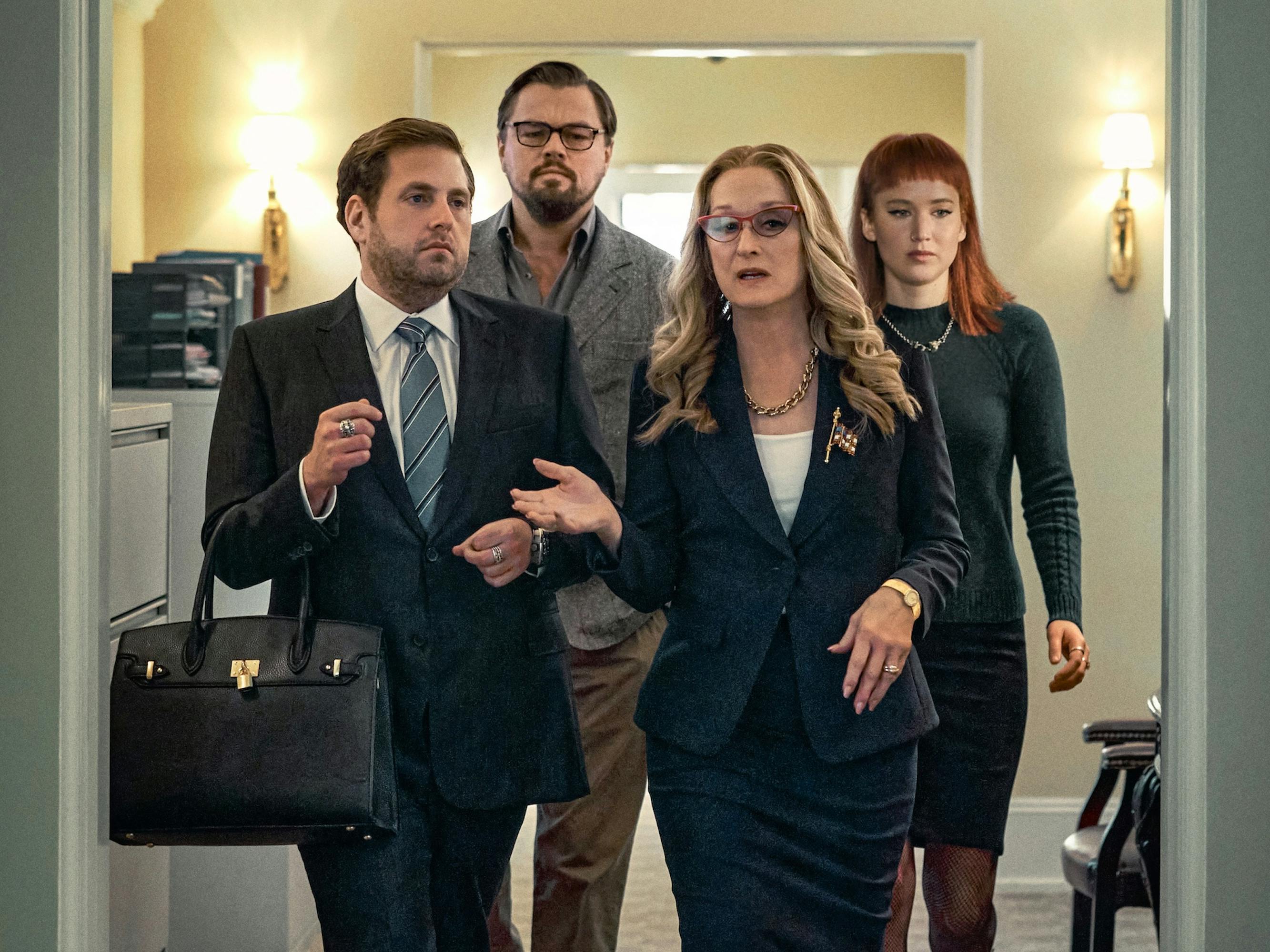 Jonah Hill, Leonardo DiCaprio, Meryl Streep, and Jennifer Lawrence walk through a White House hallway. Hill wears a navy suit and carries a black Birkin. DiCaprio wears khakis and a grey blazer. Streep wears a navy skirt suit. Lawrence wears a grey knit dress.