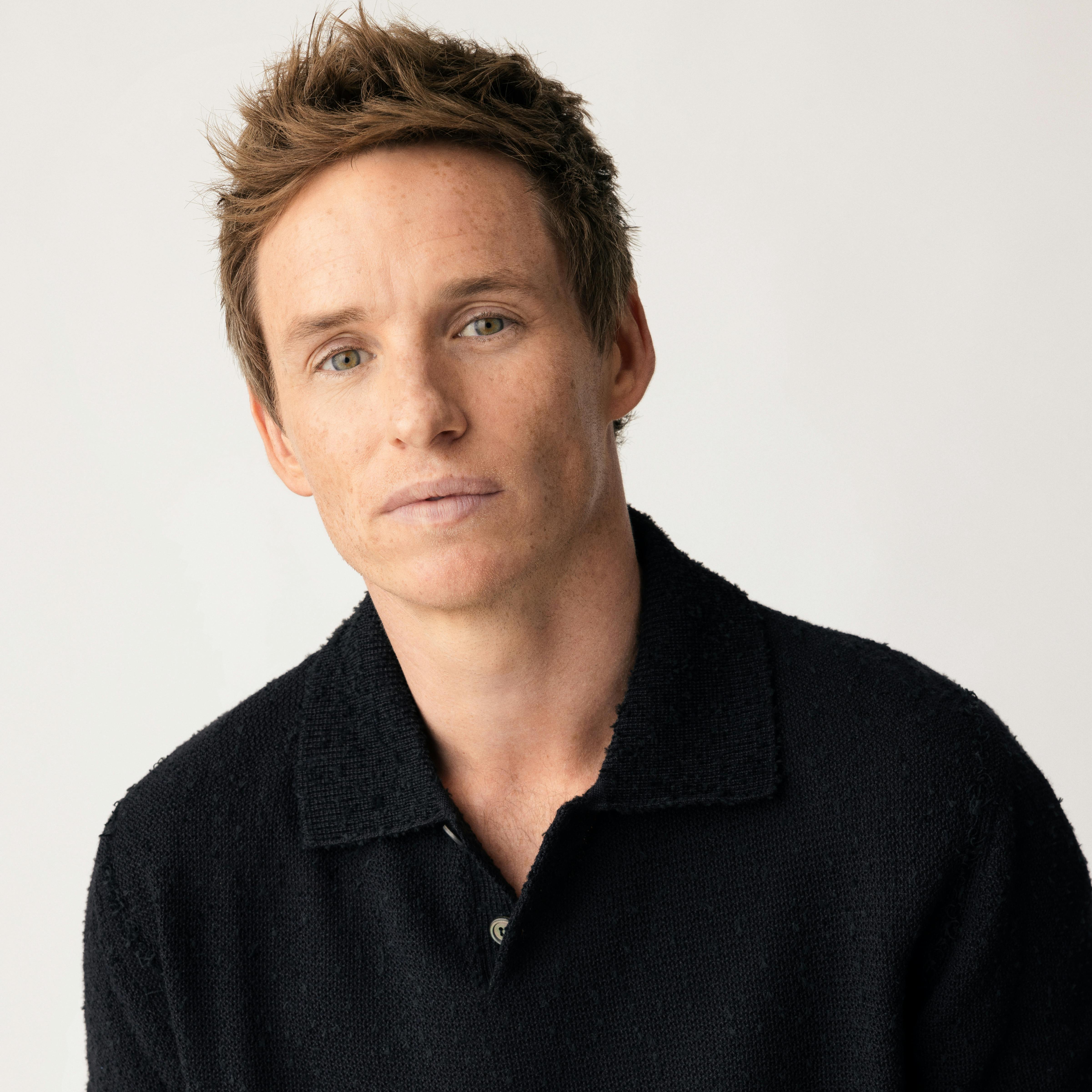 Eddie Redmayne wears a dark shirt and poses against a white background.