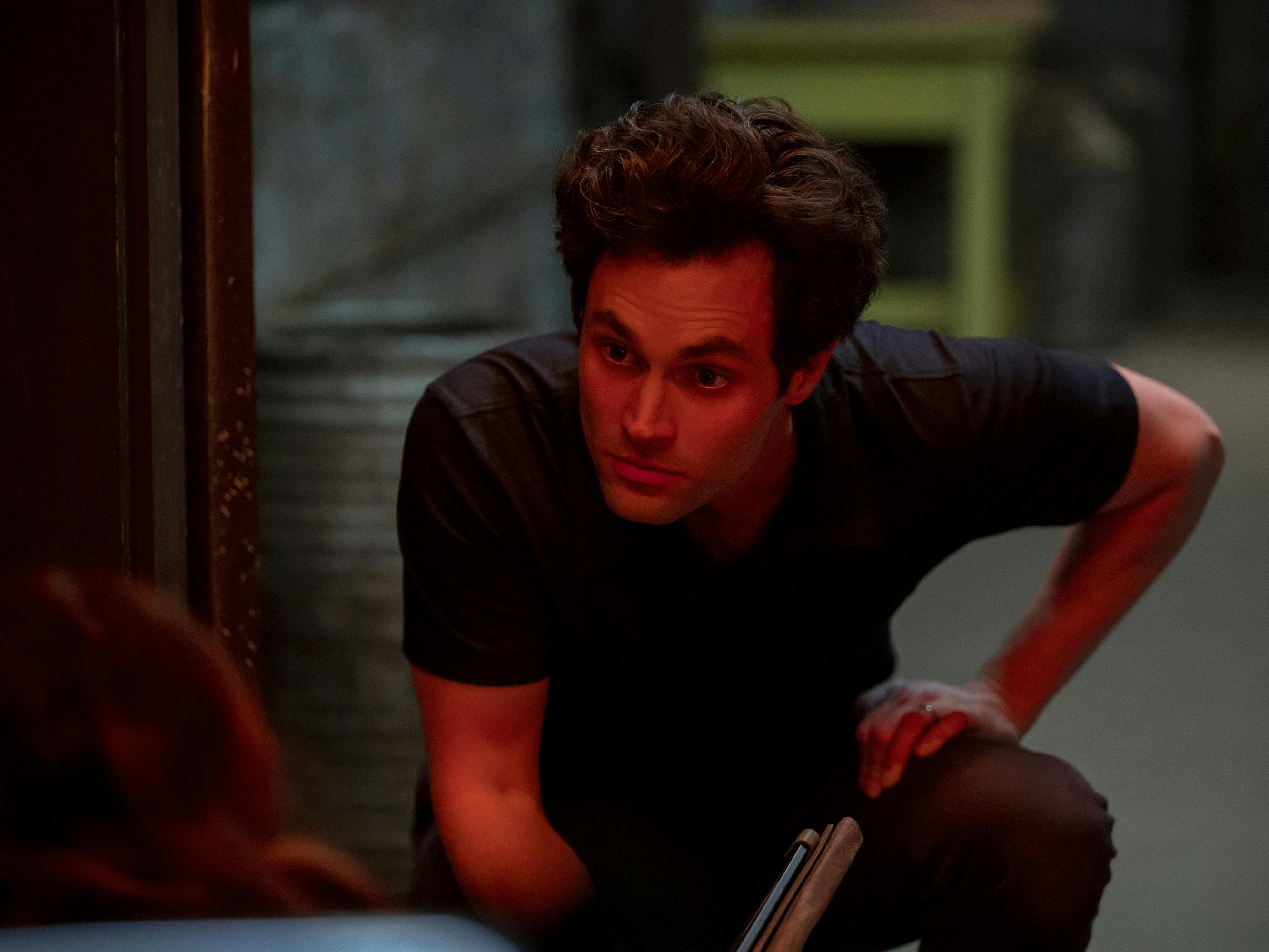 Joe Goldberg (Penn Badgley) looks threatening at someone off camera, lit by a red light.