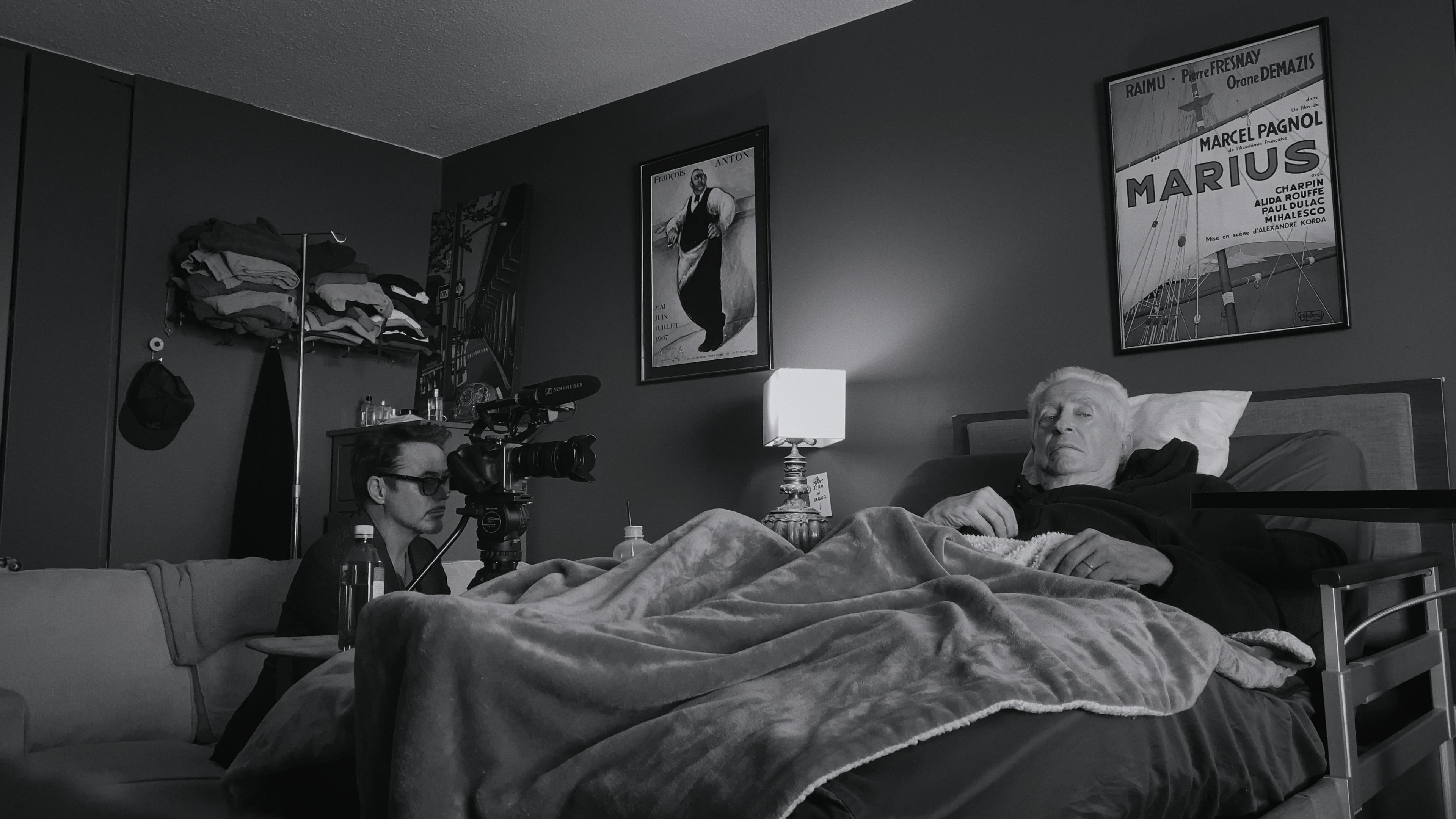 Robert Downey Jr. sits in a chair across from Robert Downey Sr.'s bed in this black-and-white picture.