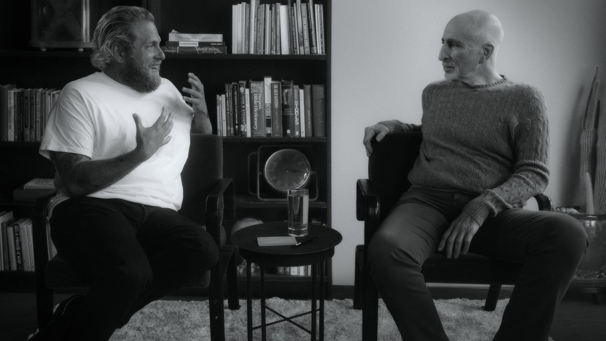 Dr. Phil Stutz wears a cable-knit sweater and sits in a library with Jonah Hill, who wears a white t-shirt and dark pants. 