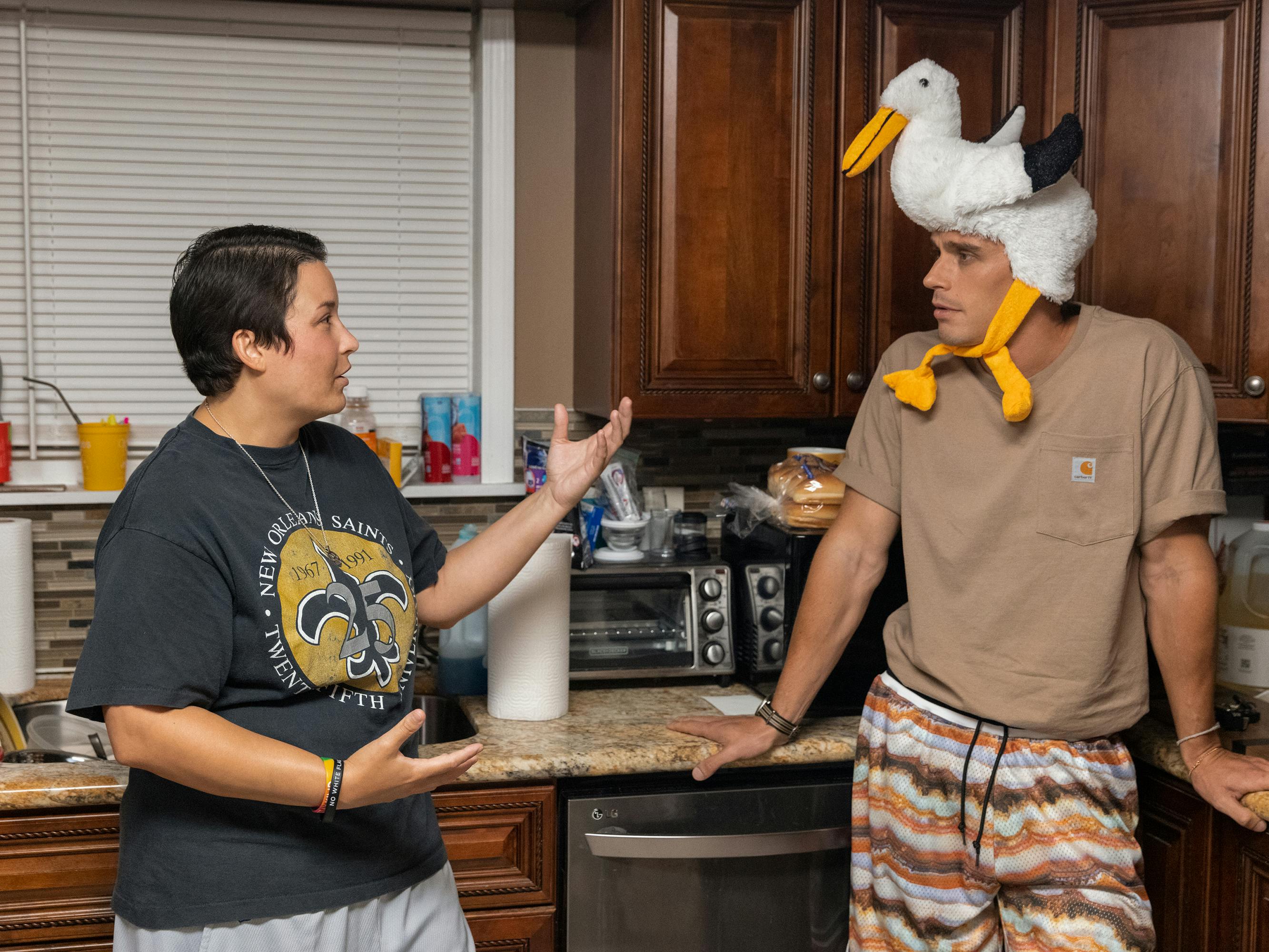 Stephanie and Antoni stand in the kitchen together. Stephanie wears a Saints shirt and grey shorts, and Antoni wears patterned shorts and a Carhartt t-shirt. His most notable sartorial choice is the seagull hat he wears. I wonder what the story behind that is.
